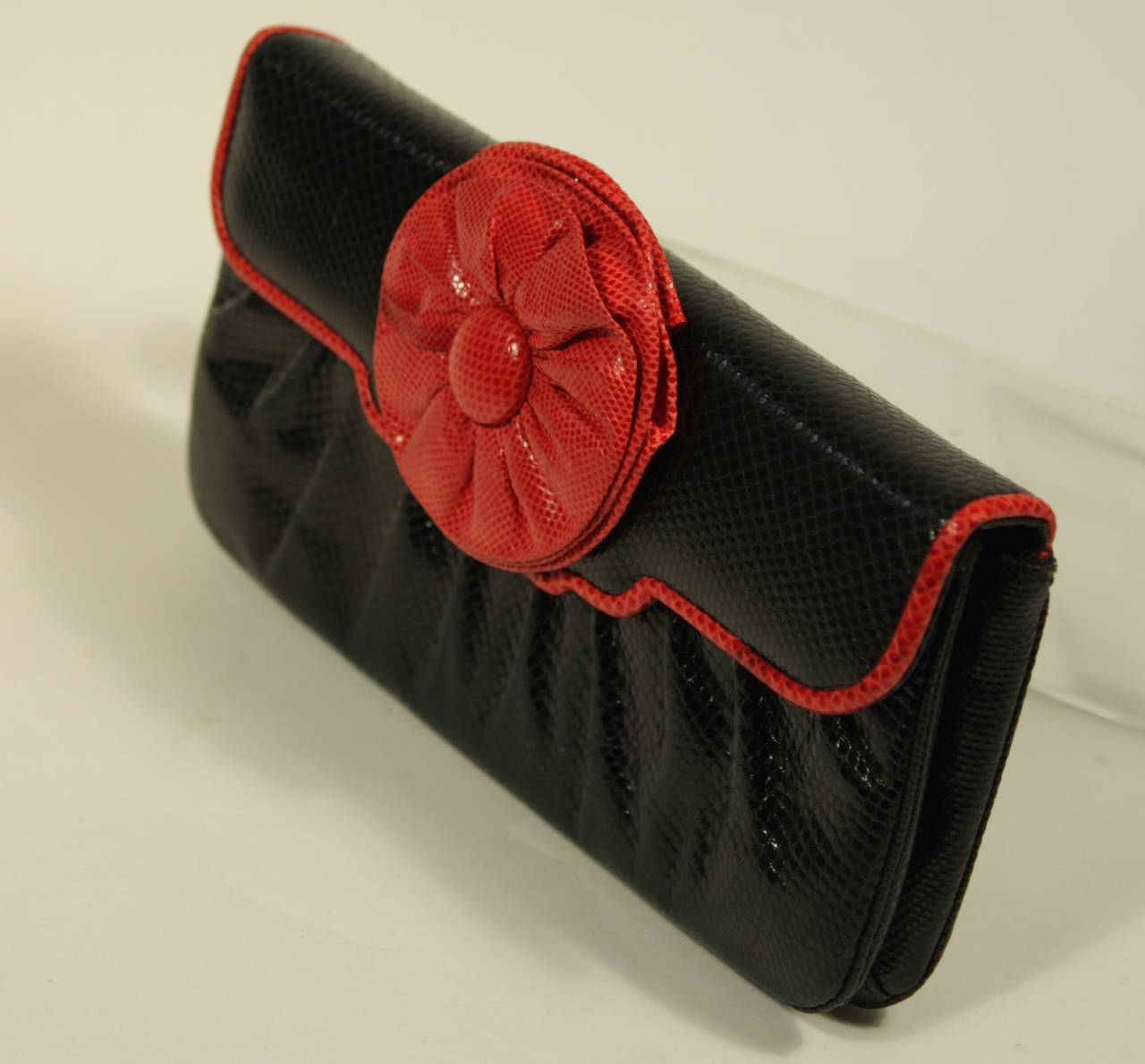 This Judith Leiber design is available for viewing at our Beverly Hills Boutique. The clutch is composed of a black snakeskin trimmed with red. There is a ruffle flower detail and magnetic snap closure. The silk interior is in pristine condition and