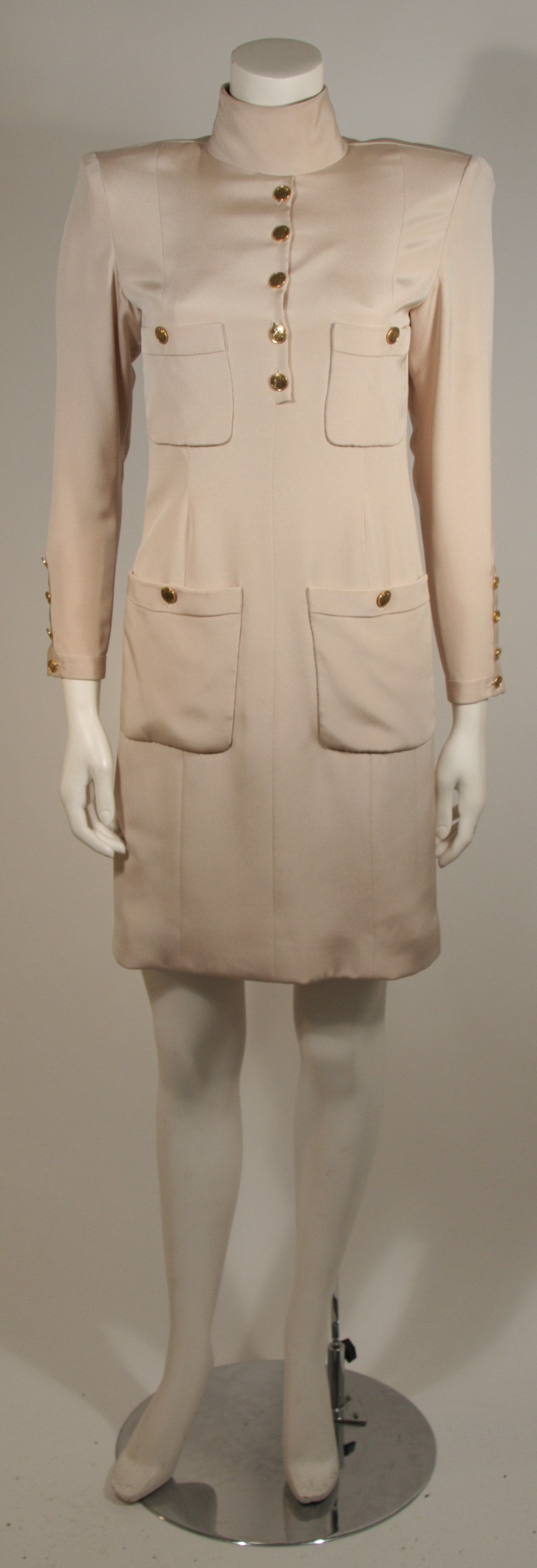 This Chanel design is available for viewing at our Beverly Hills Boutique. The dress is composed of a champagne silk and features four front pockets with gold button details. There is a center back zipper for ease of access. The item does not have a