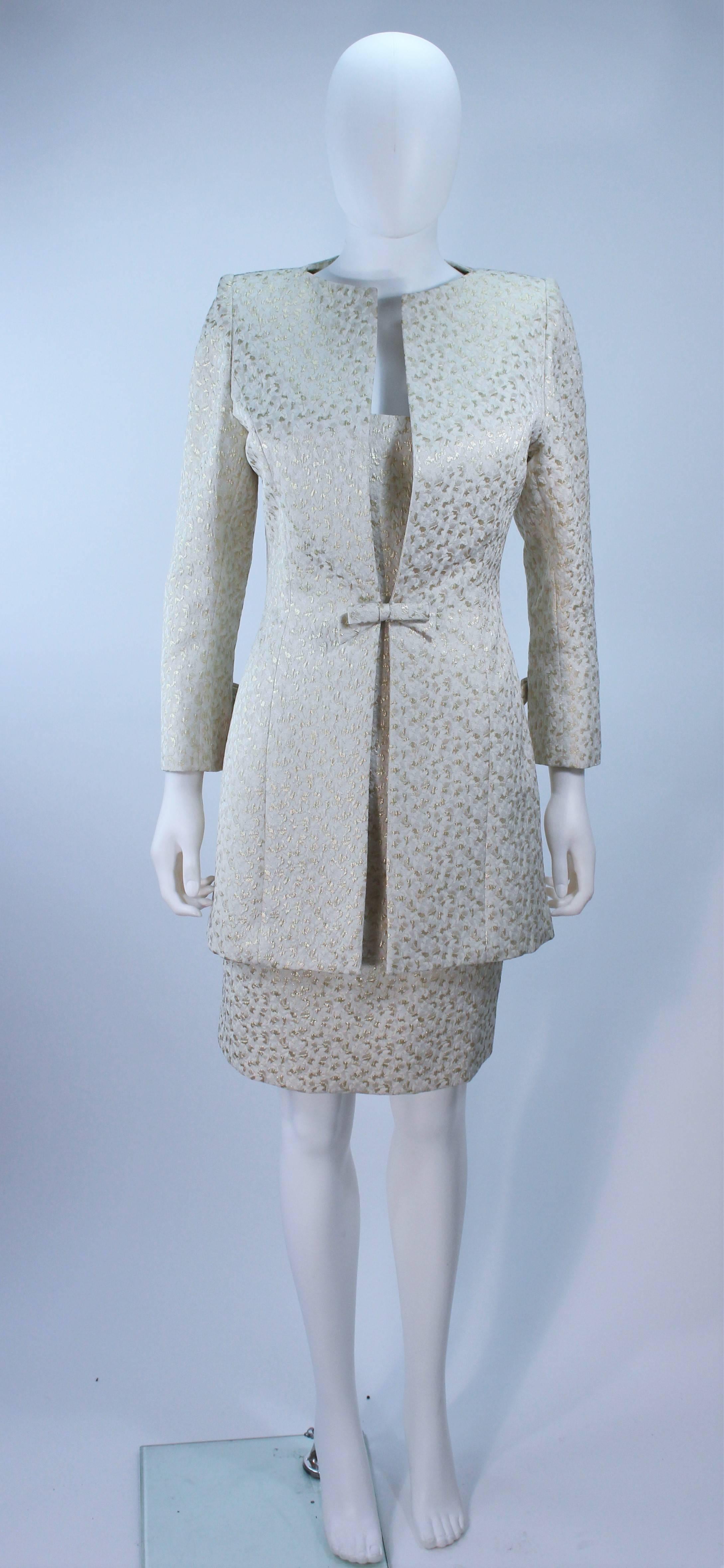 This Travilla  ensemble is composed of an off white and gold metallic brocade silk. The coat features bow accents at the center front and sleeves with a hook and eye closure. The dress features a classic silhouette with a zipper closure. In