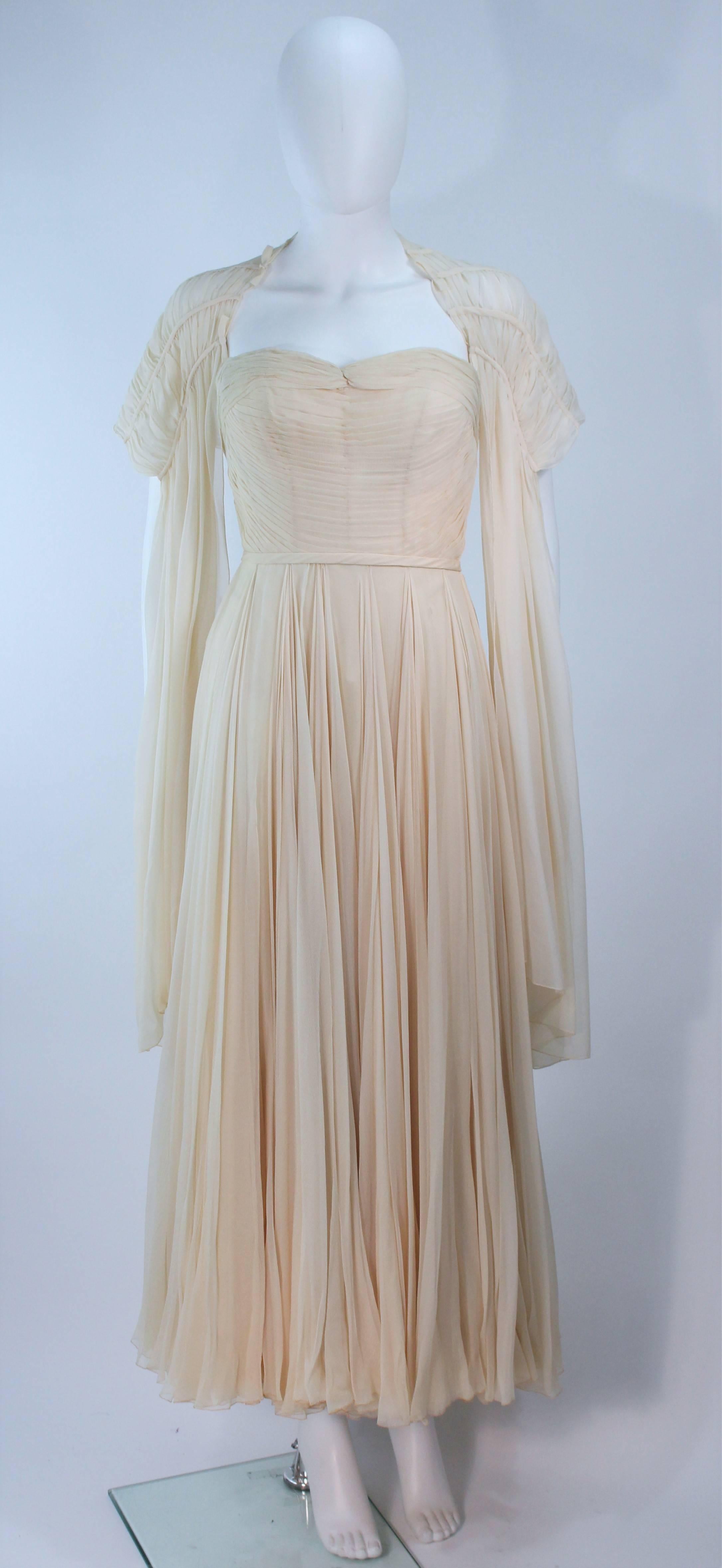 This gown and wrap are composed of an off white, cream hued silk. Features a gathered bodice with pleated full skirt. There is interior boning and zipper closures. The wrap has a gathered draped design with bow accents. In excellent vintage