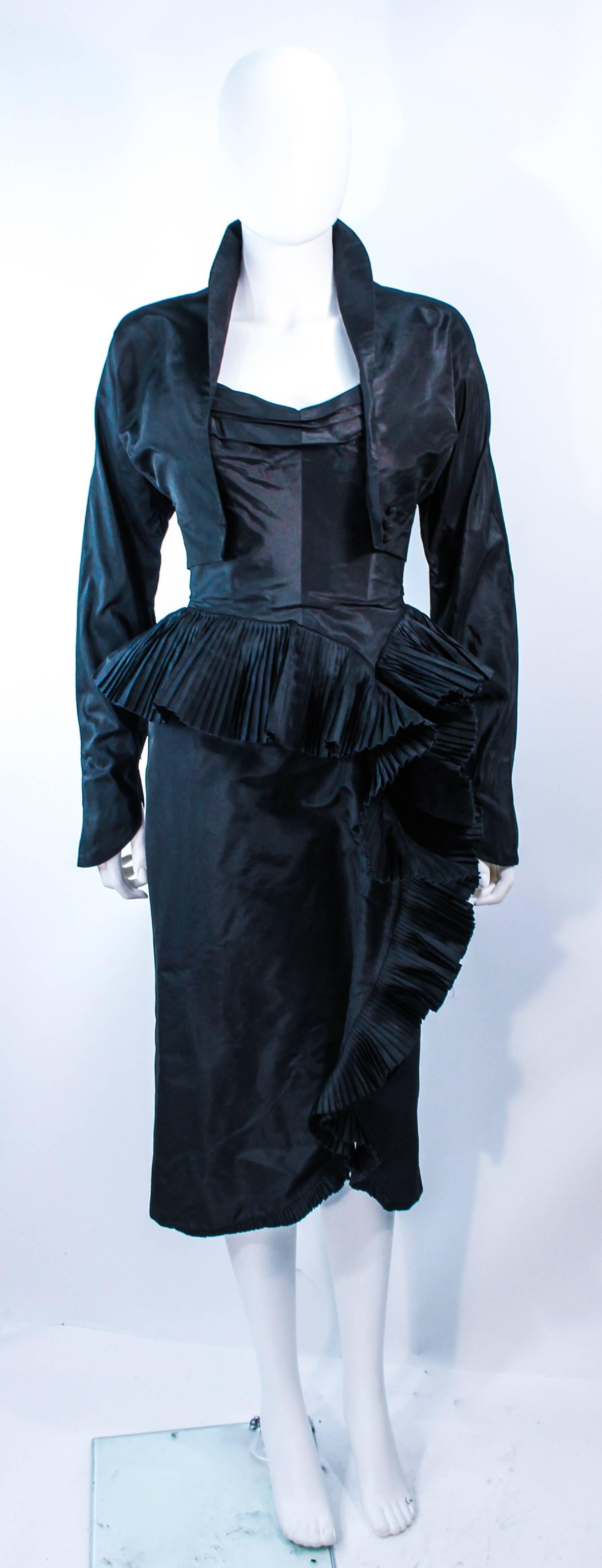 This Irene ensemble is composed of black silk. The strapless cocktail dress features a pleated bust with side draped, cascading ruffle, with interior boning. The jacket features a cropped style with front buttons. In excellent vintage