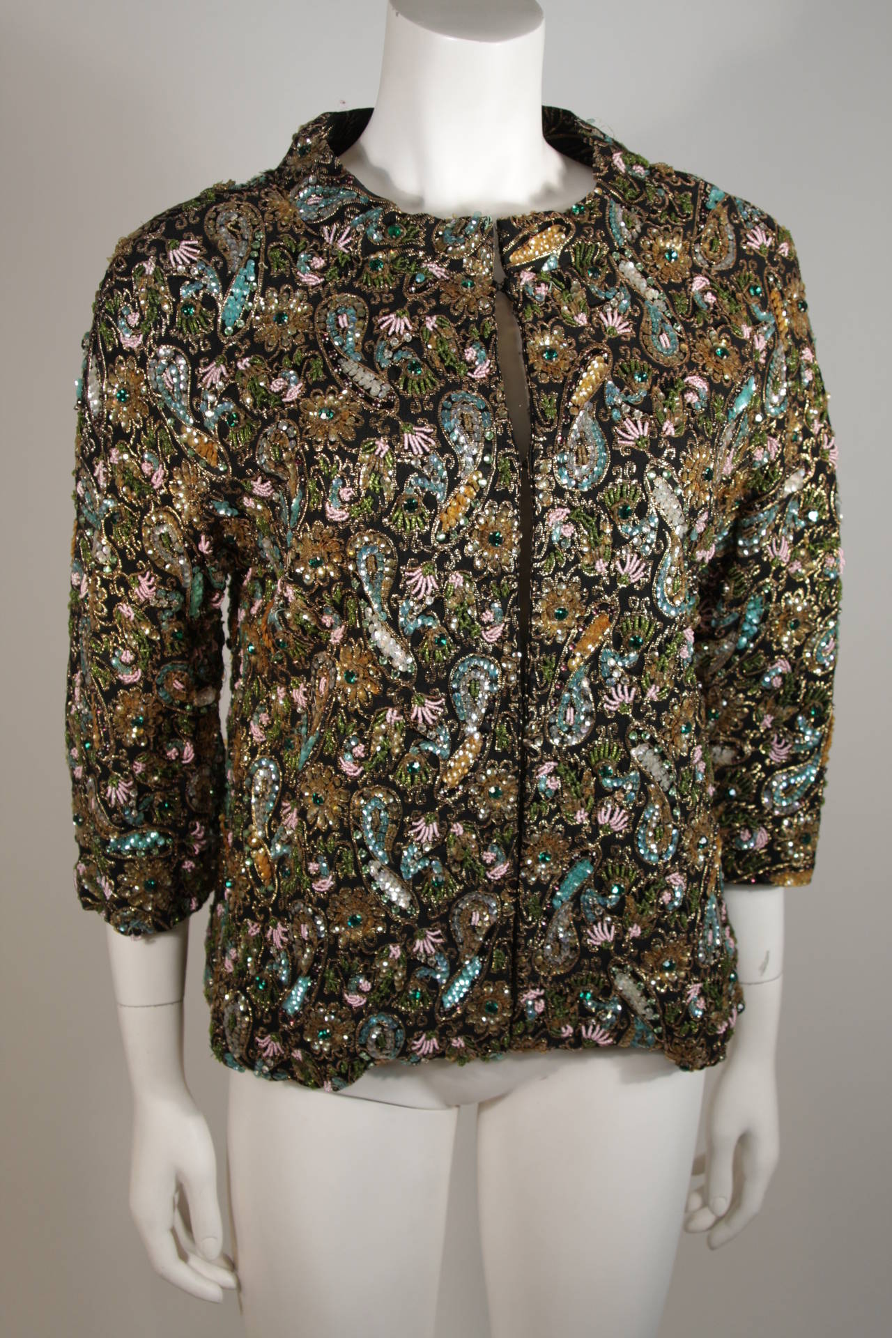 This beautifully embellished Diane Imports jacket features a heavily beaded floral motif in aqua, emerald, pink, black, and gold beads. Composed of a rayon, silk, and metallic blend. There are center front hook and eye closures. This is a stunning