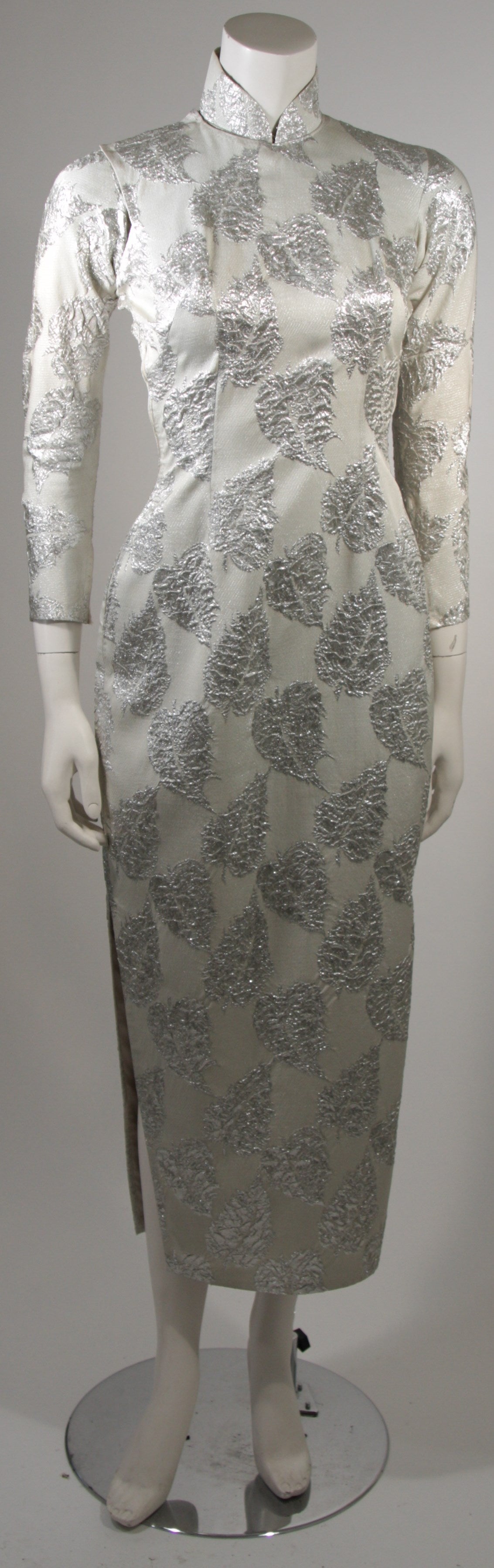 This vintage Asian inspired custom made cheongsam cocktail dress is composed of a silver silk brocade and features a mandarin styled collar, a body skimming design with slits at the side for ease of movement. There is a side zipper and snap