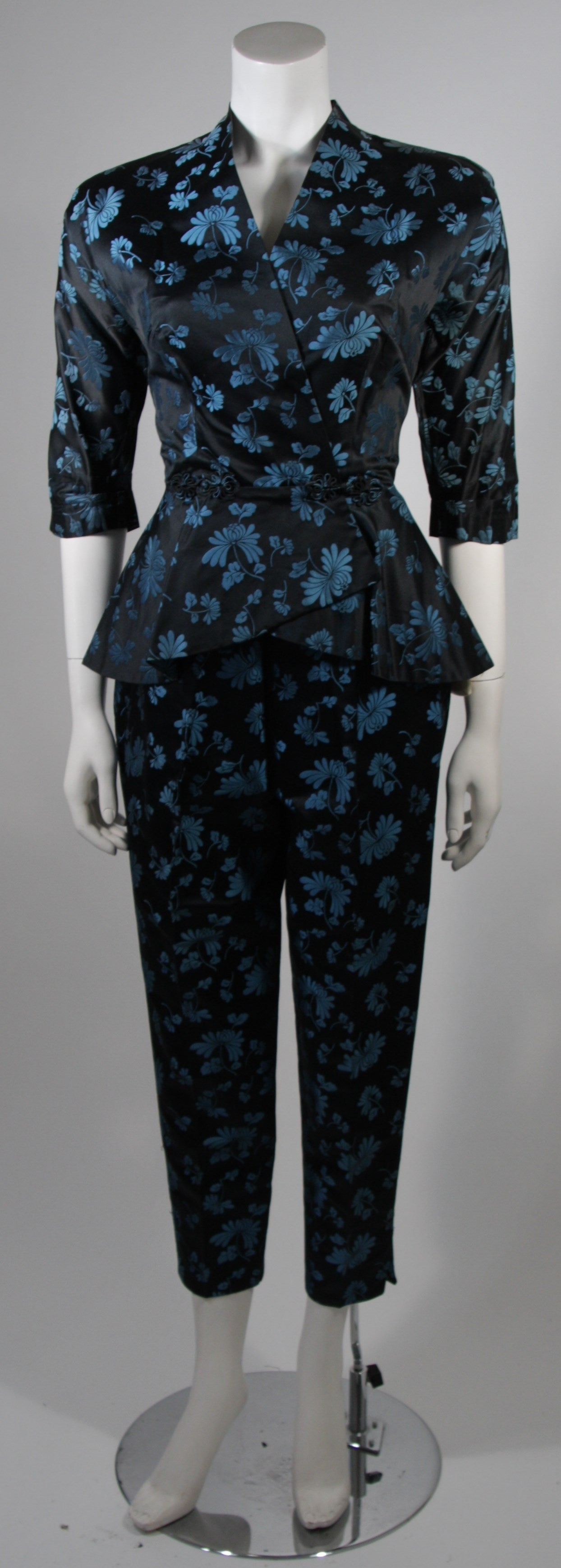 This Dynasty pant suit is composed of a black and blue silk with an Asian inspired floral motif. The jacket features a peplum style, elbow length sleeves, and center front closures. The pants are a classic cigarette style falling just above the