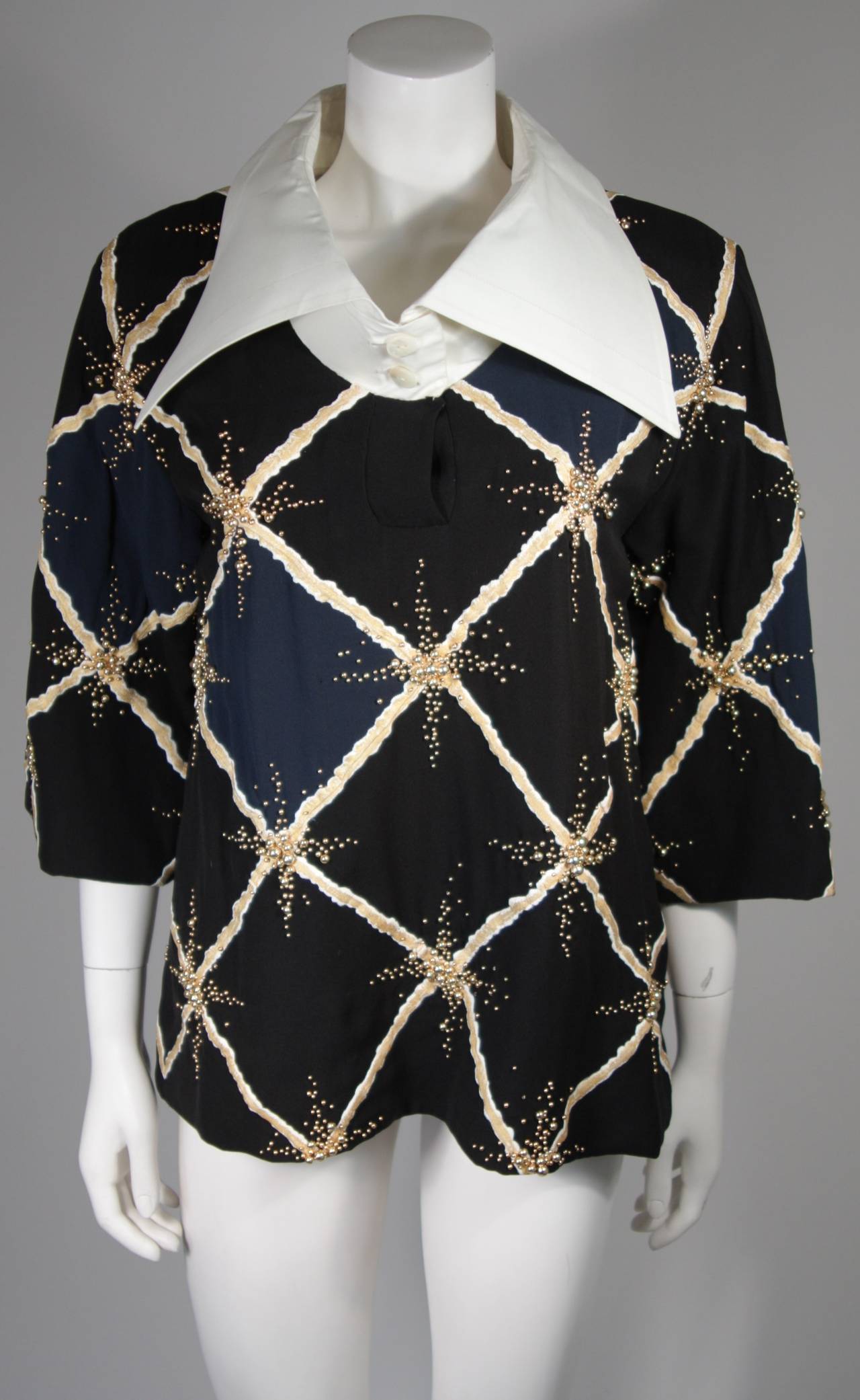 This Pierre Balmain blouse is composed of a navy, black, and white patterned silk with embellishments. The linen collar is fused for a three dimensional effect, which causes it to sit up right. The embellishments are small gold beads. In excellent