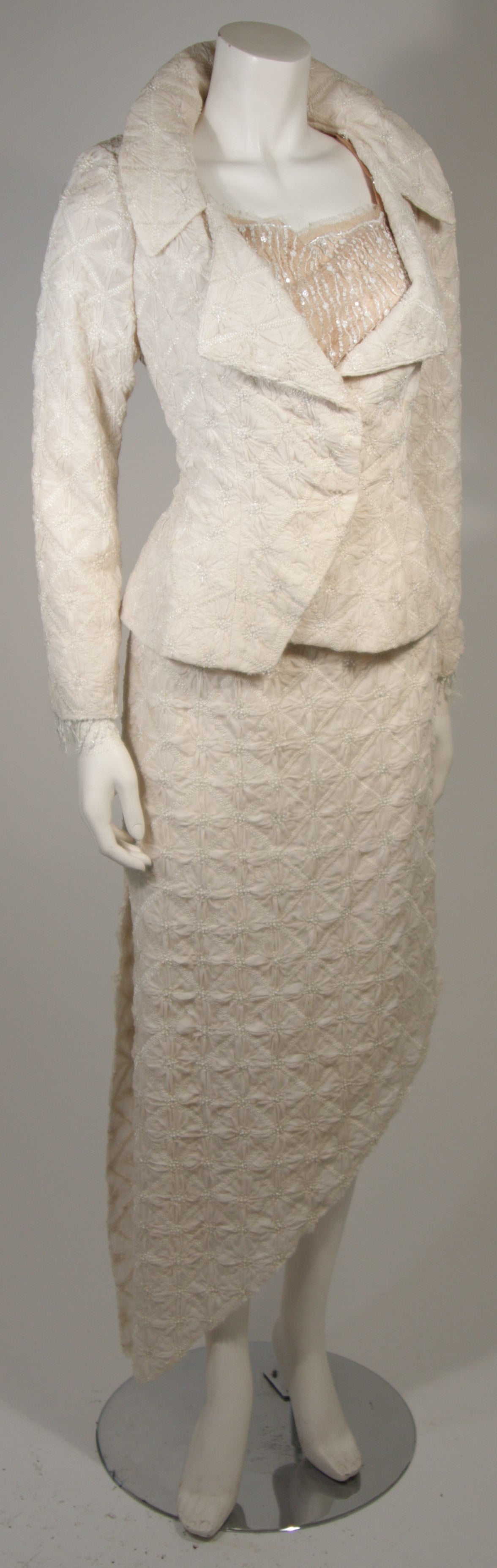 This Nolan Miller attributed ensemble is composed of off white silks and adorned with small pearl embellishments. The jacket features a large collar and a flare at the waist, there are center front snap closures. The skirt has an asymmetrical design