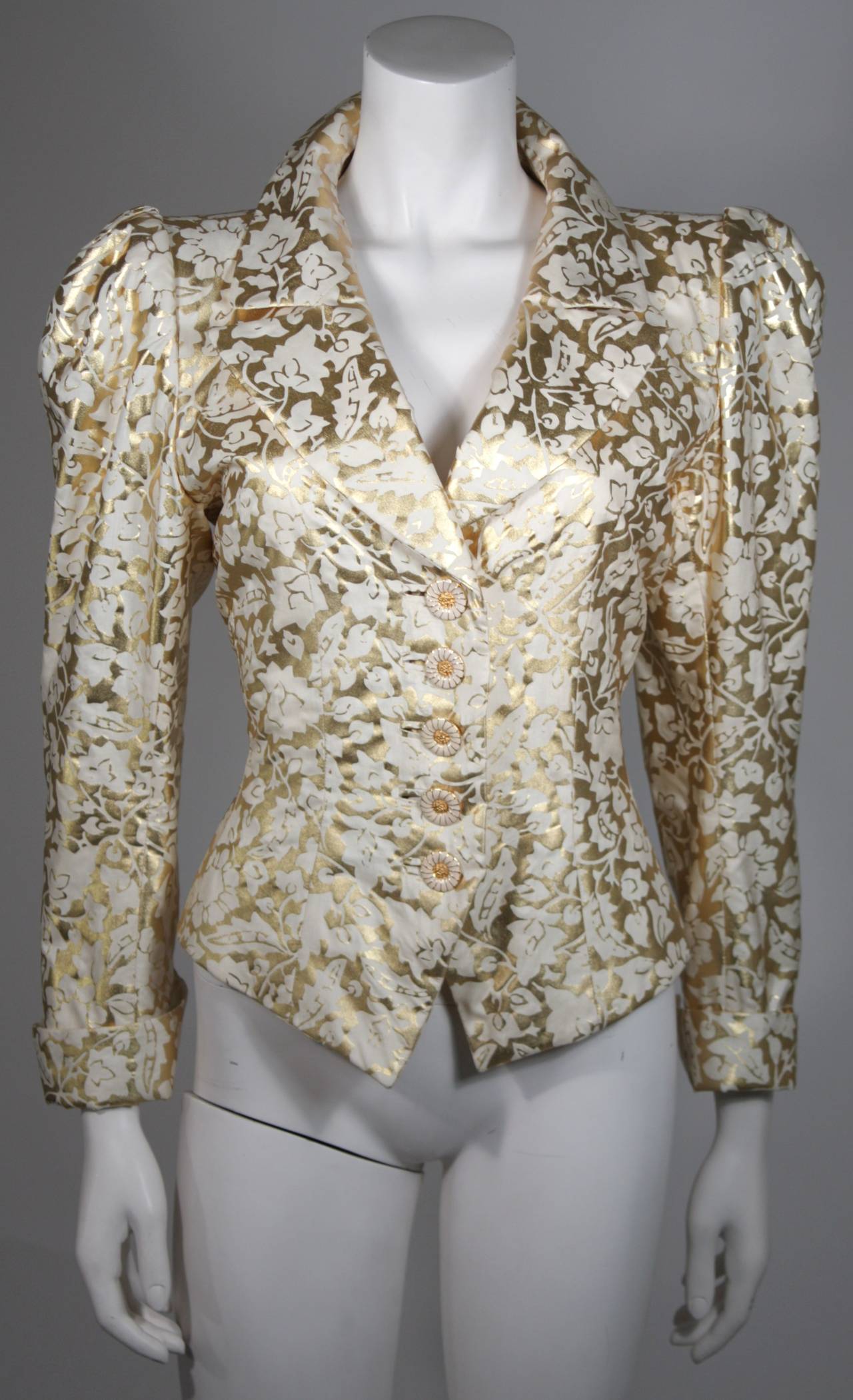 This Yves Saint Laurent jacket is composed of a gold and white fabric with a floral motif. The sleeves feature a puff style with cuff. There are gold with white enamel daisy style buttons at the center front. In excellent vintage condition. Made in