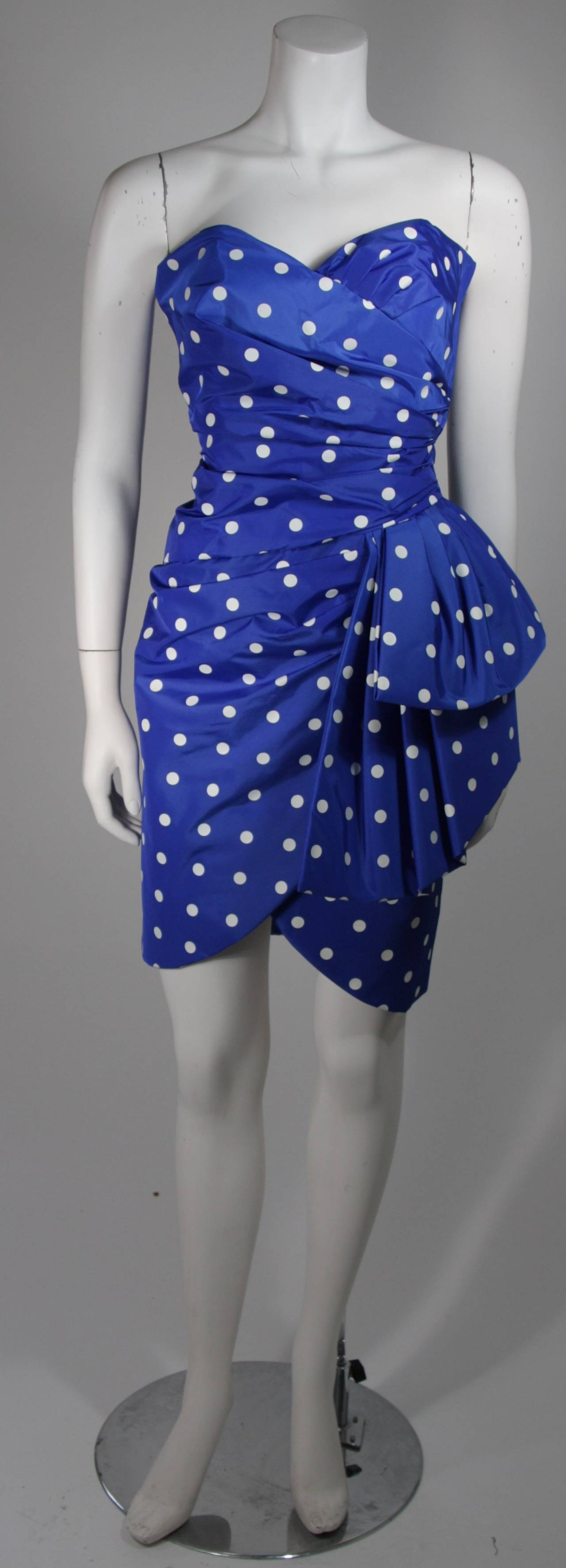 This Victor Costa cocktail dress is composed of a royal blue and white polka fabric. The gathered design is accented by a large side draping bow at the hip. There is a side zipper closure. In excellent condition. Made in United States Of America.