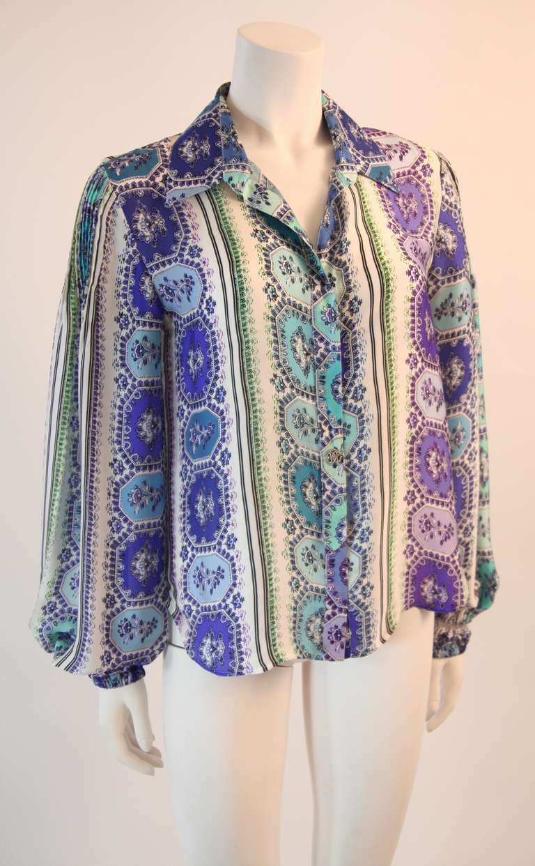 This is a wonderfully flowing white silk Roberto Cavalli blouse
with a beautiful purple, turquoise, black, and green print. The blouse has a spread collar,logo rhinestone button detail placket, high low hem, and blouson sleeves with a fitted