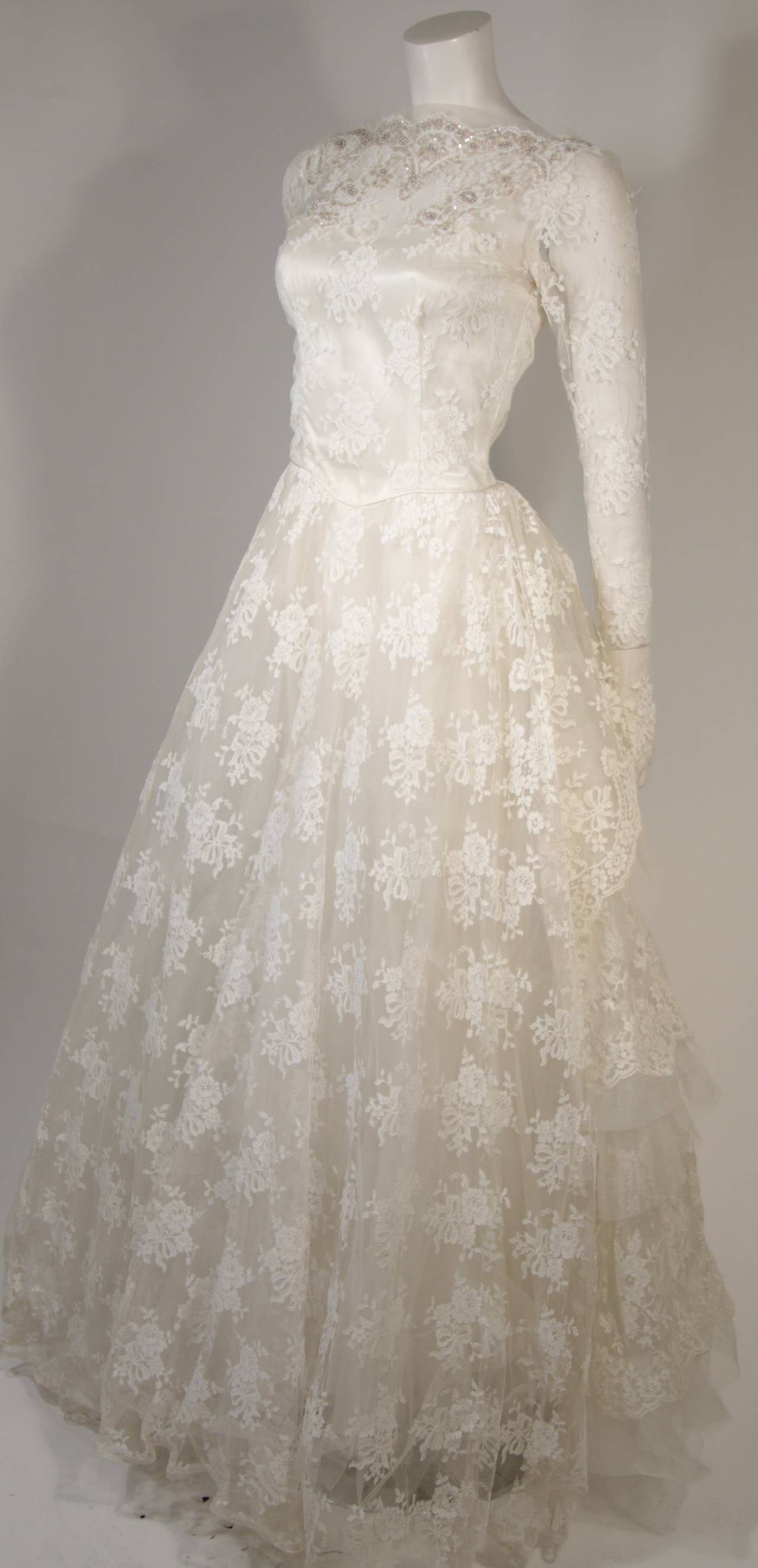 This vintage wedding gown is fabricated from layers of white lace featuring scalloped edges and satin. The scalloped neckline is adorned with sequins and pearl accents. There are center back button closures. The dress has some slight discoloration