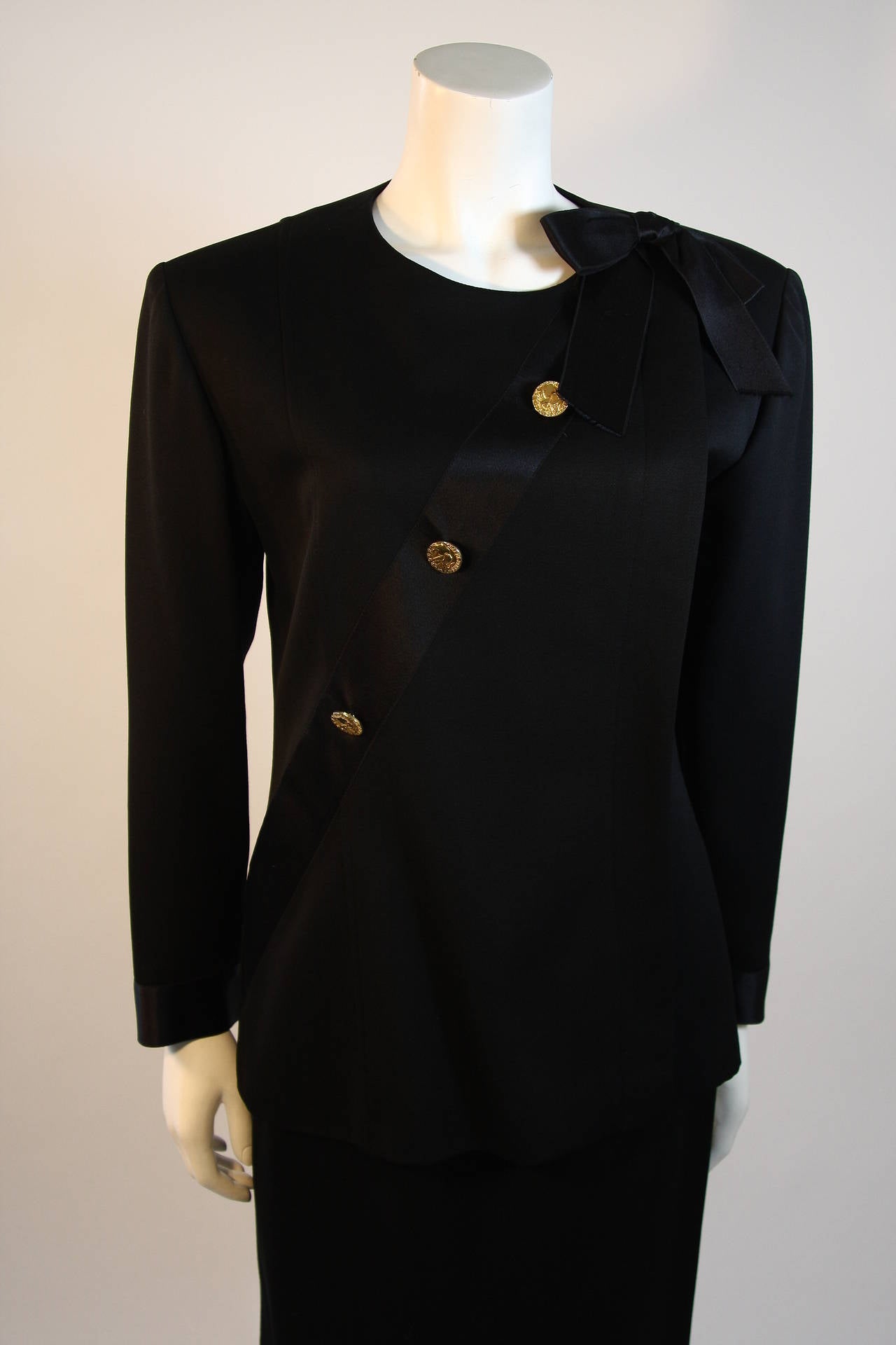 Black wool jacket with hidden button placket at left side, with cross body black silk satin ribbon adorned with gold elephant embossed Chanel Paris buttons, finished with a bow at the left shoulder. The cuffs of the jacket are trimmed in same 1.75