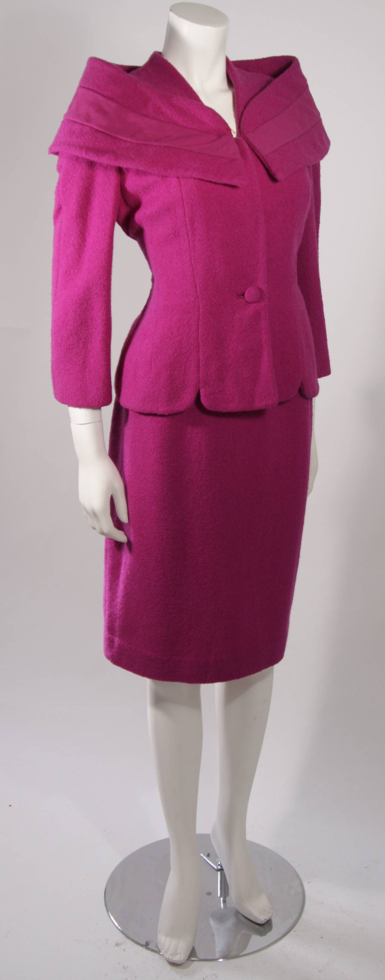 This is a Lilli Ann ensemble. The suit is composed of a purple wool. The jacket has center front closures and features a wrap style accent at the shoulders. The skirt is a fitted pencil silhouette and has a zipper closure. 

Measures