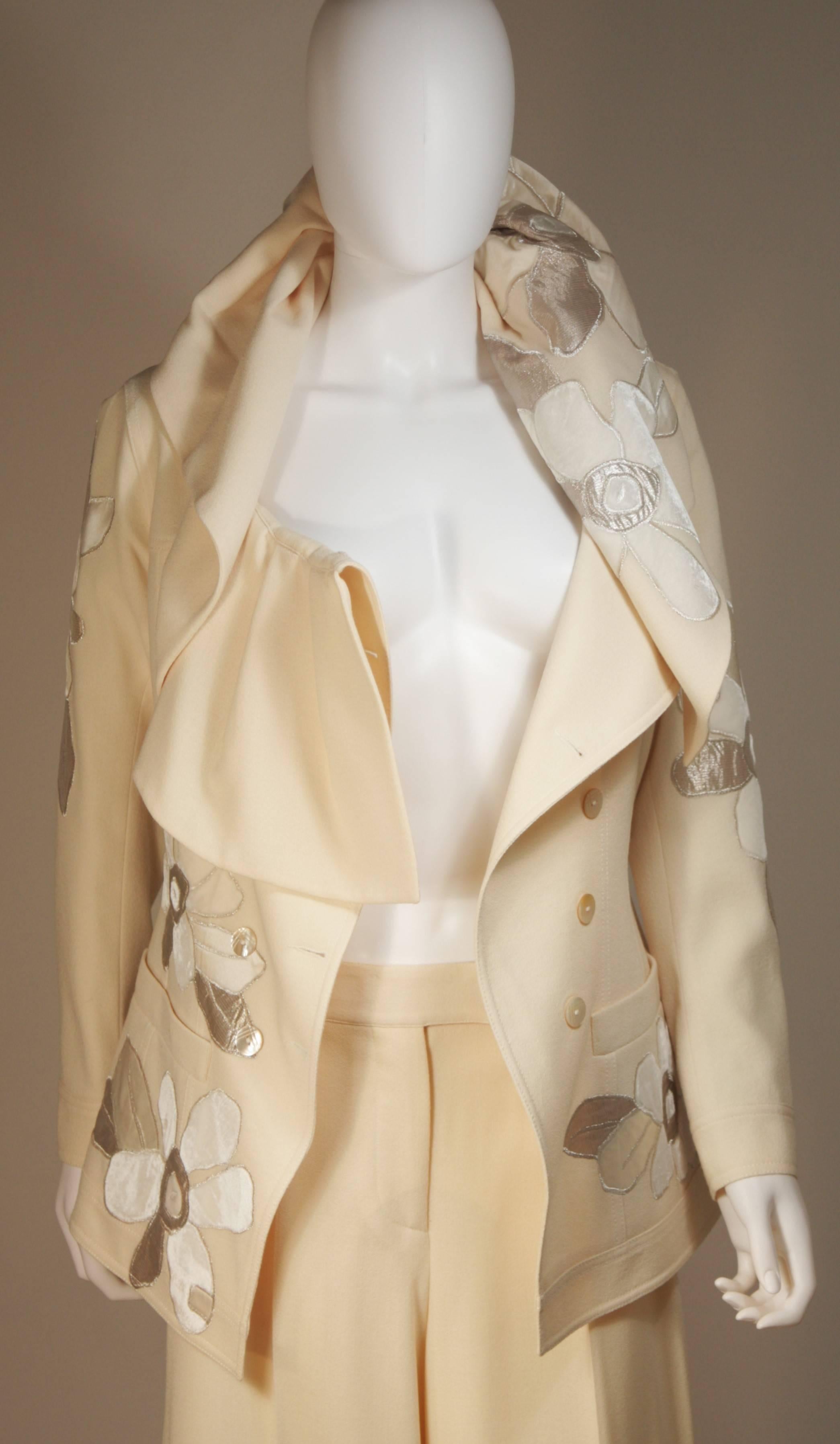 This John Galliano suit is composed of an off white fabric with silver metallic applique. The suit features a dramatic ruffled neckline which can be styled in a variety of ways. The pants are a classic wide leg style with center front crease and