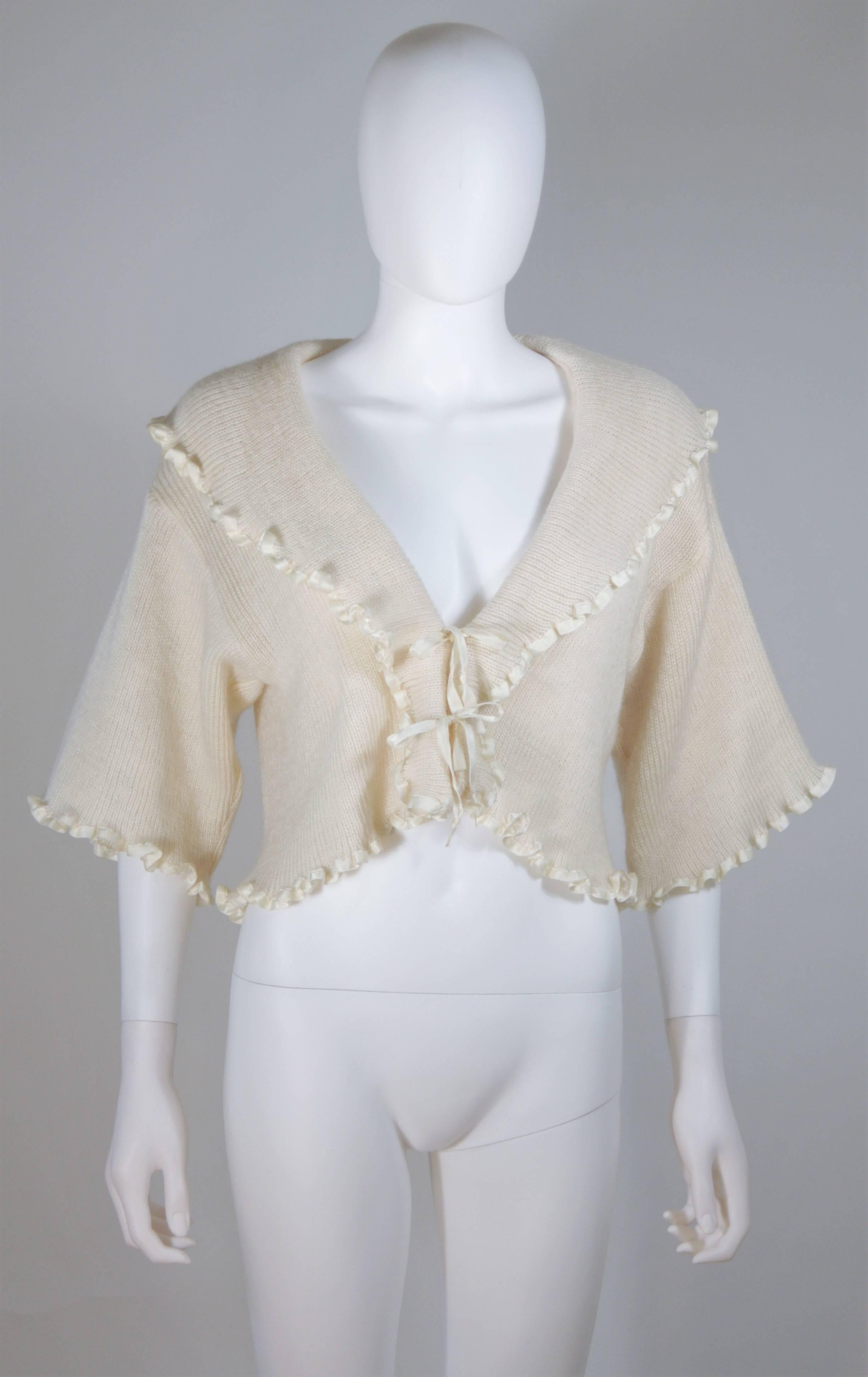 Dieser Pullover von Christian Dior ist aus einer cremefarbenen Kaschmirmischung gestrickt. Es hat einen gerüschten Saum mit Satinbesatz und wird vorne mit einer Krawatte geschlossen. In ausgezeichnetem Vintage-Zustand. 

  **Bitte vergleichen Sie