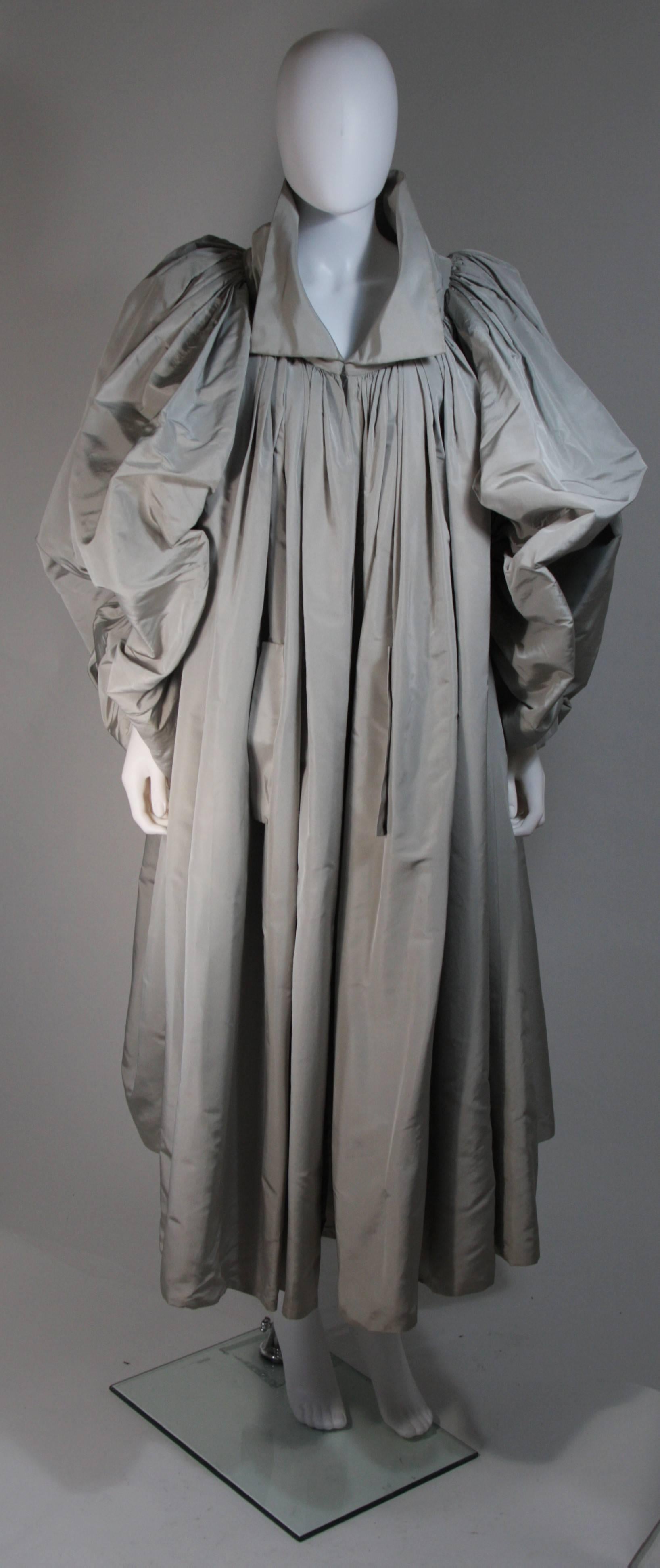 This Galanos design is composed of a grey silk. The dramatic design features a hook and eye closure at the collar end. There is gathering at the neckline and rounded puff style sleeves. This coat is sold in 