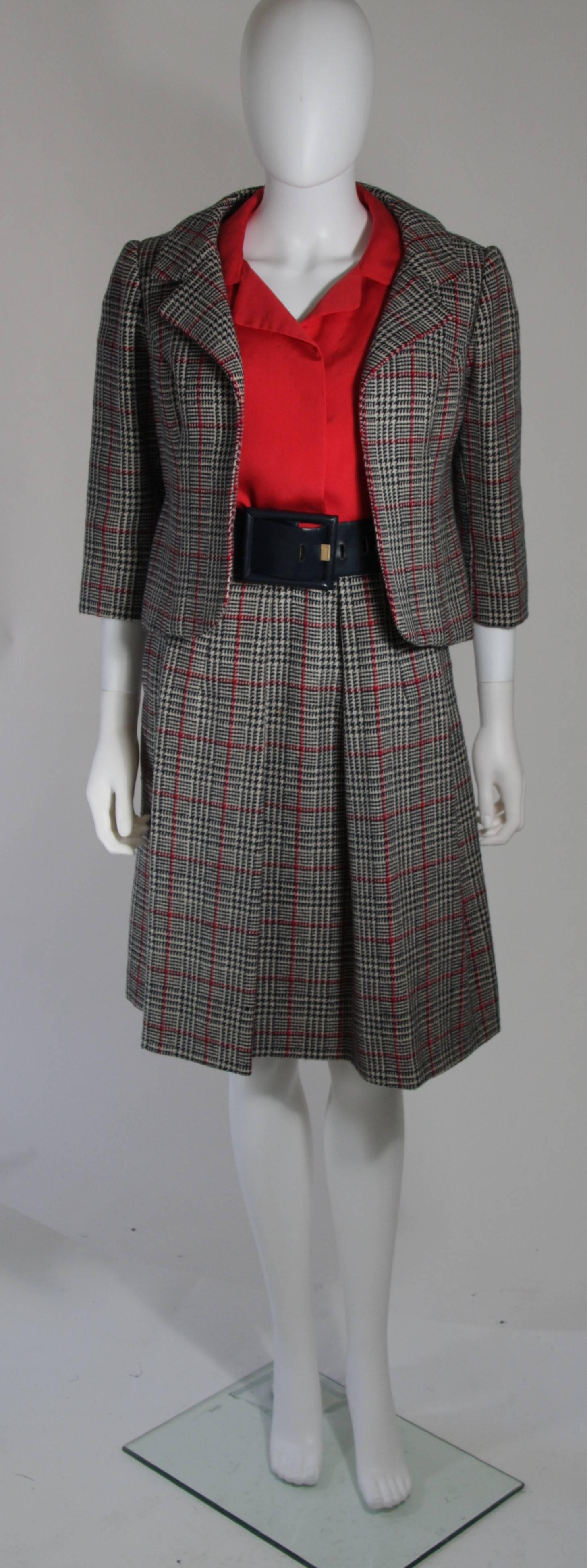 This Galanos skirt suit is composed of a plaid wool in red, white, and black. The blouse (designed by Dorothy Veeder) is composed of a red silk and has center front snap closures. The jacket has an open style. The skirt features a pleated front and