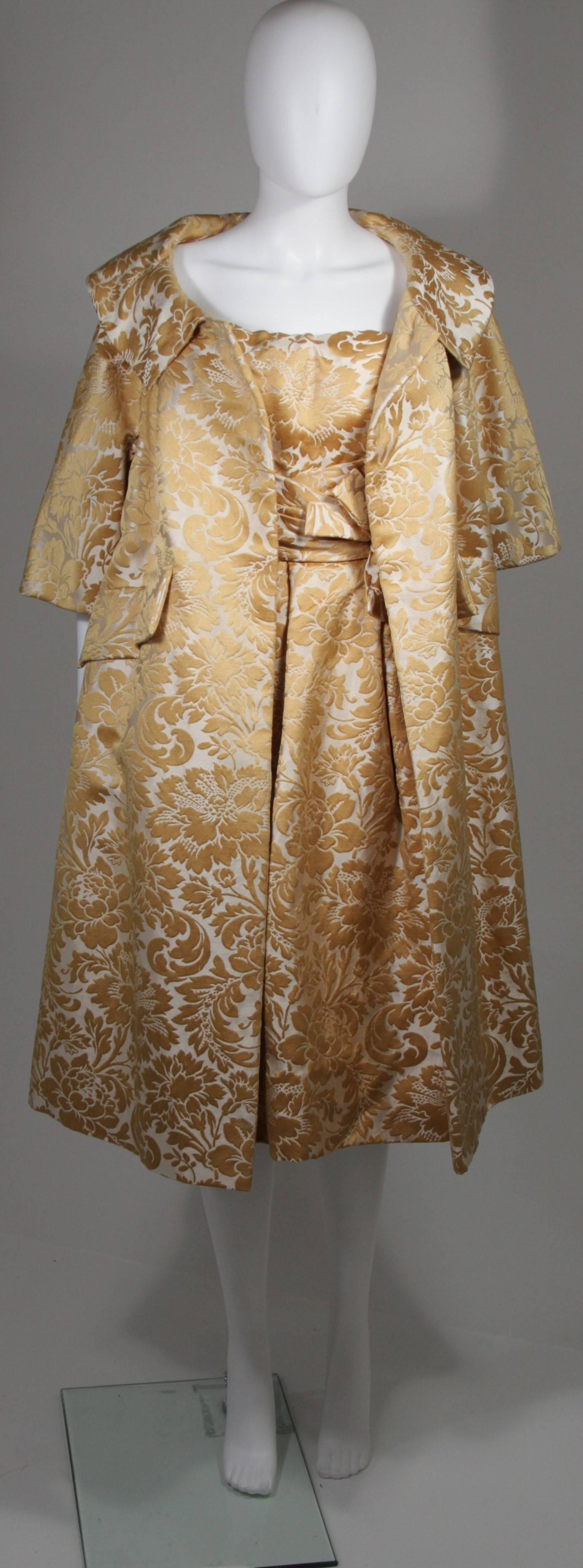 This Samuel Winston evening ensemble is composed of a gold and cream brocade. The dress features a draped style at the waist which culminates to a bow. There is a zipper closure. The large opera coat features a button closure with hook and eye. In