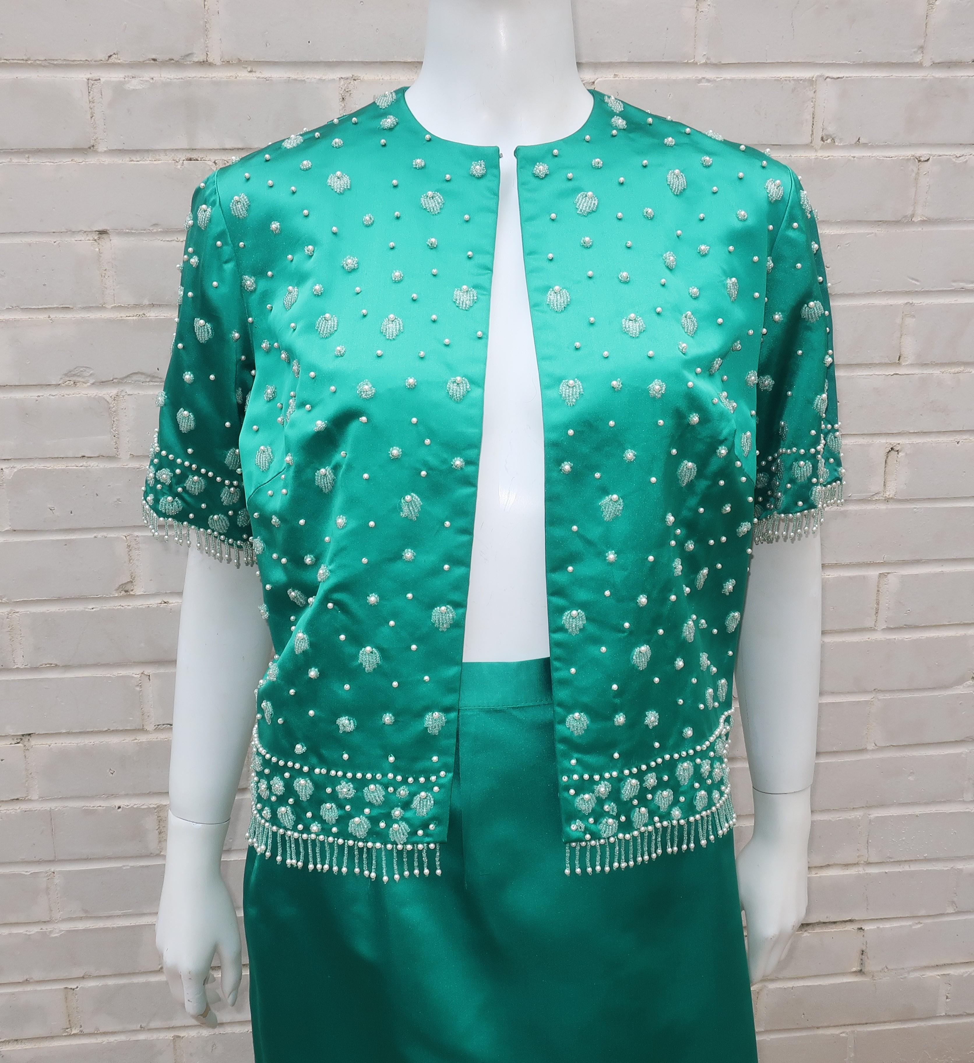 Exotic elegance exudes from this C.1960 jade green satin cropped top jacket and skirt ensemble.  The silk and rayon blend is fully lined and bears a bluish hint to the rich green shade offset by pearls and seed beads in a floral design with fringe