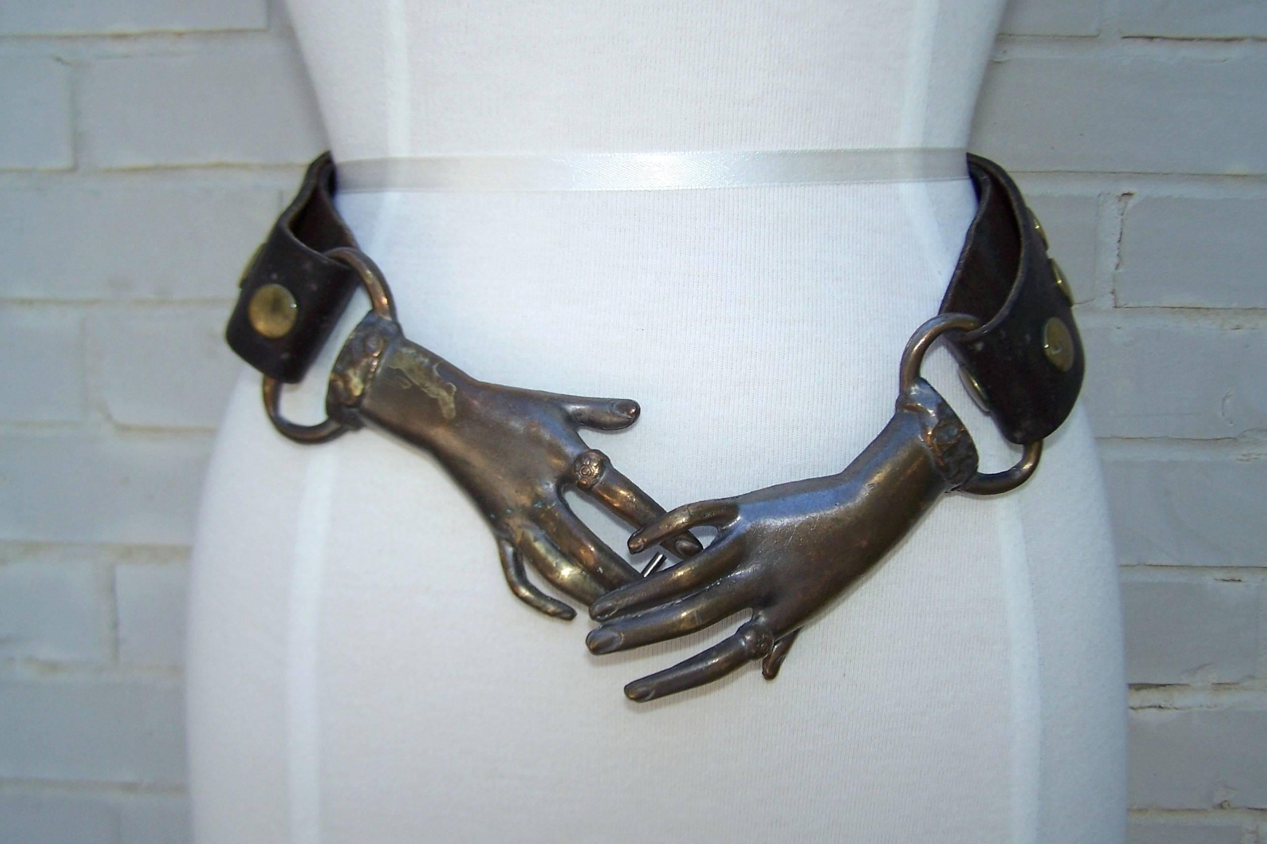 Extra hands have long been a favorite motif for fashion designers over the years including Elsa Schiaparelli.  This 1970s version is a pair of Victorian style clasping metal hands with an antiqued brass finish supporting a chunky dark brown leather