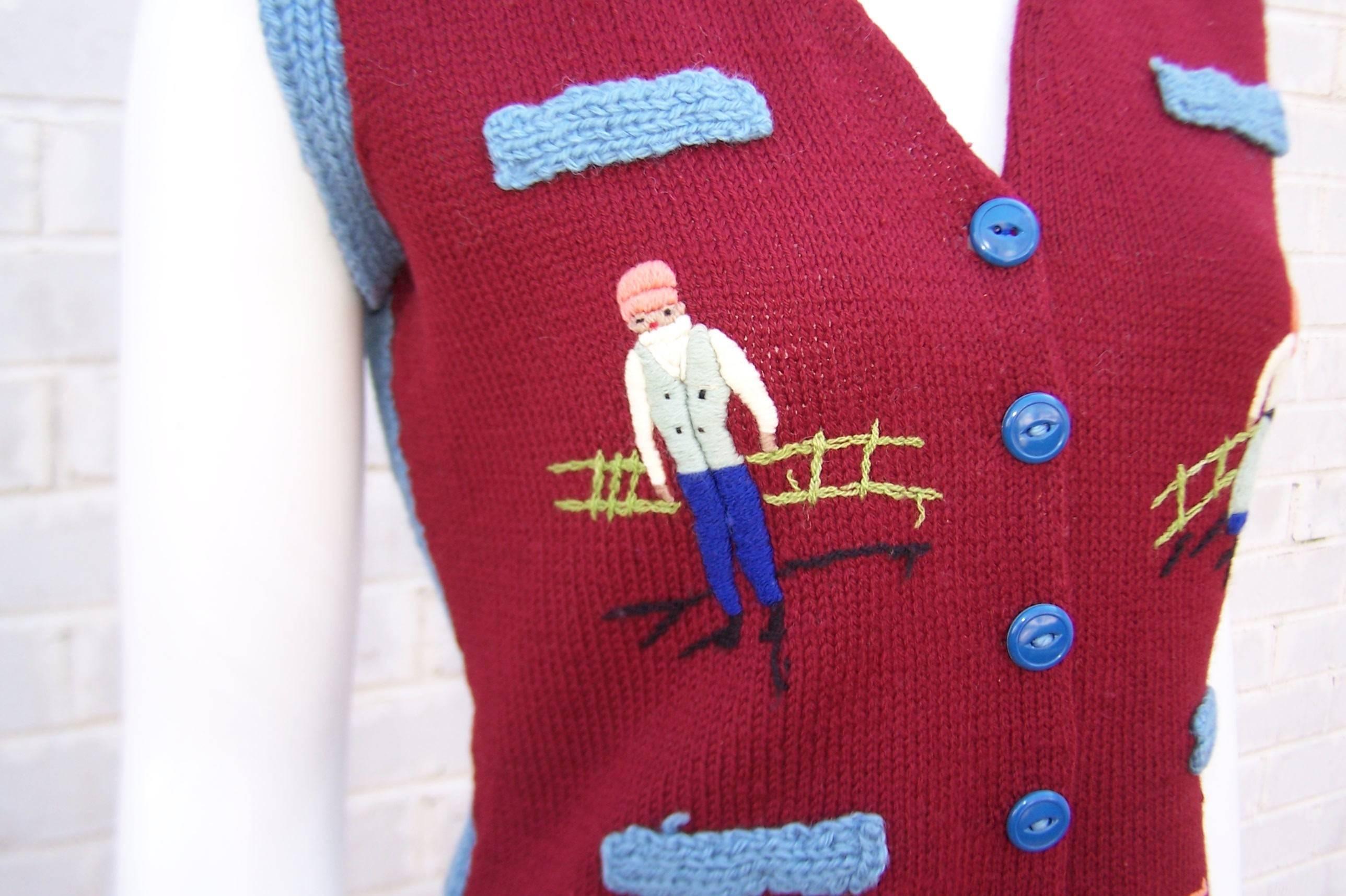 This C.1970 hand knit sweater vest is decorated with charming imagery .  The vest is styled like a menswear waistcoat with buttons at the front and faux pockets.  It is a burgundy red at the front and baby blue at the back with folk art depictions