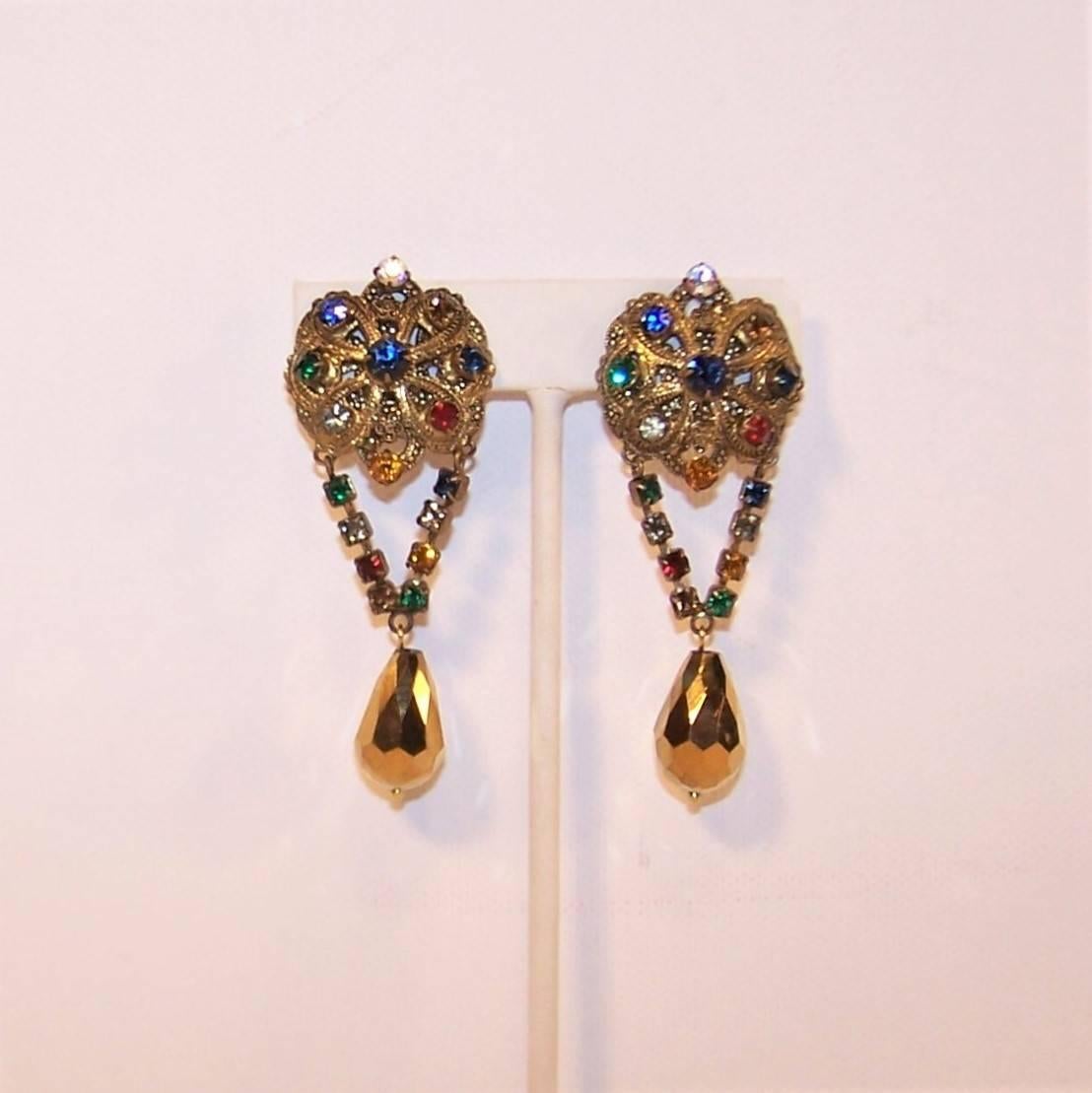 These 1950's clip on earrings have an old world style with regal details including an antiqued gold metal finish and jewel tone rhinestones.  The filigree base is reminiscent of Miriam Haskell fittings with a rhinestone chain supporting a glass