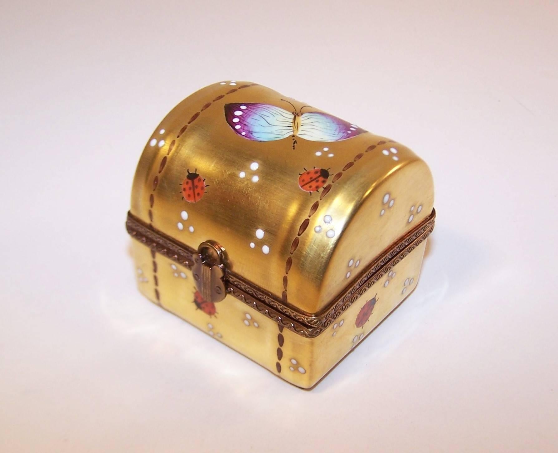 This French limoges porcelain trinket box resembles a miniature treasure chest complete with a padlock closure and faux stitched details.  It is charmingly hand painted with a butterfly and lady bugs on a rich golden background.  The interior opens