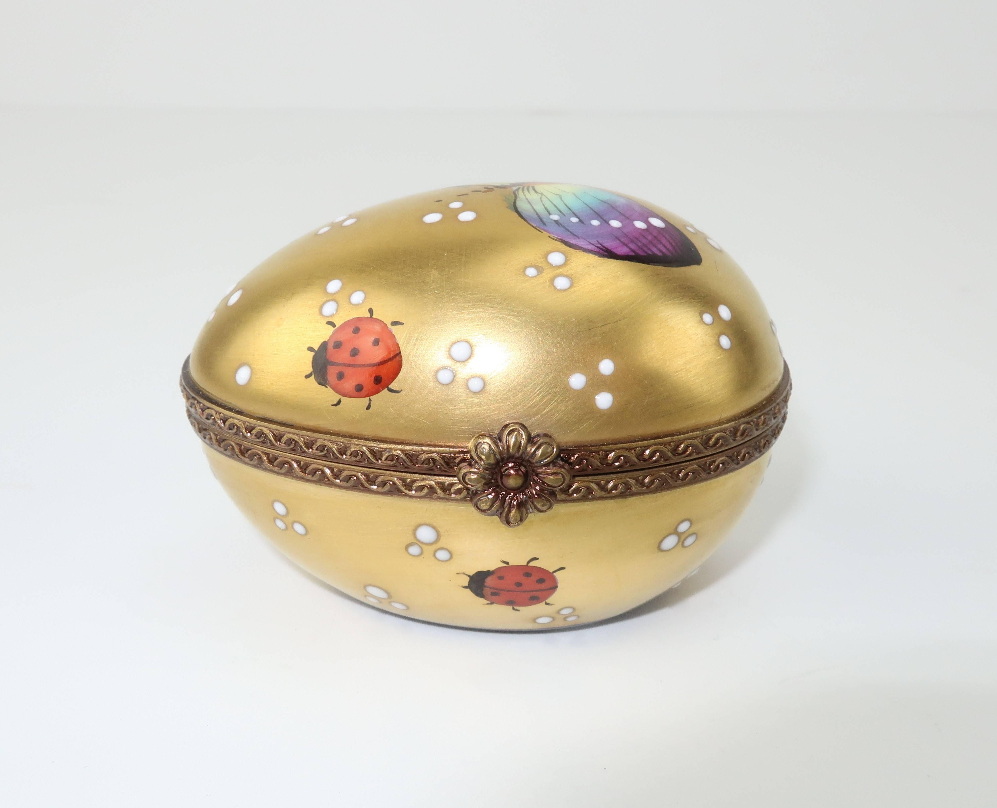 This French limoges porcelain trinket box is a delicate egg shape with a floral decorated closure.  It is charmingly hand painted with a butterfly and lady bugs on a rich golden background.  The interior hinges opens to reveal a leaf painted on the