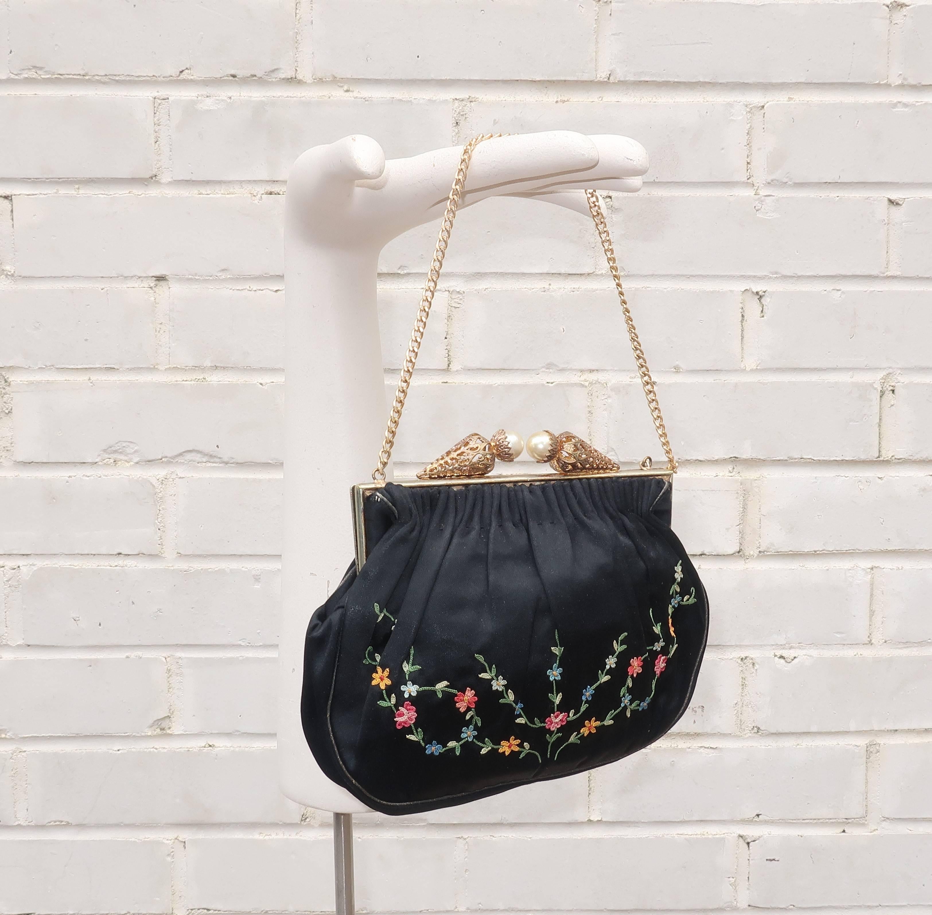 This delicate and ultra feminine 1950’s French black satin handbag features a bright floral chain stitch pattern in shades of pink, yellow, blue and green.  The gold tone frame is accented with filigreed cones topped by faux pearls which twist to