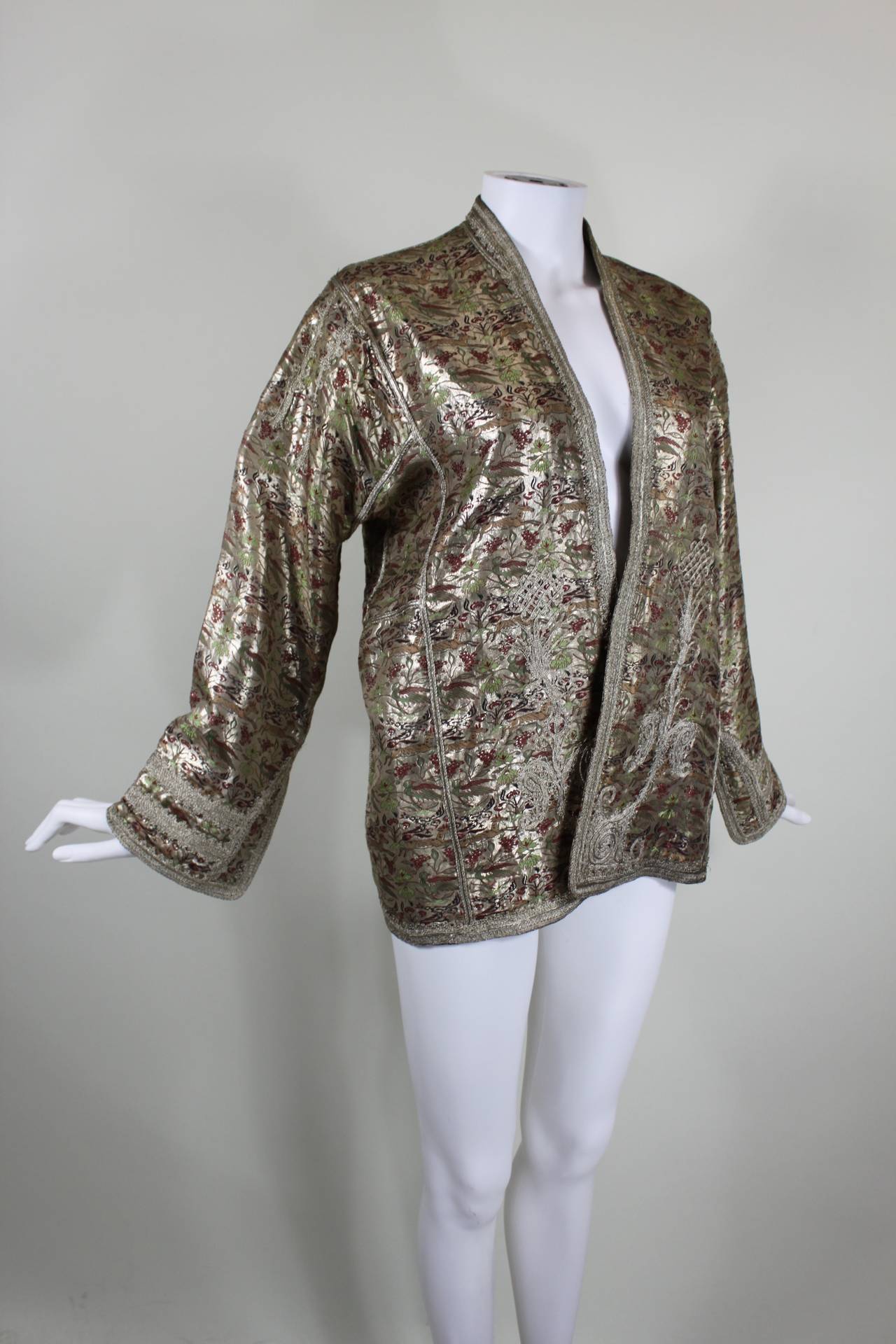 This is an absolutely stunning silver metallic jacket from the 1930s. A gazelle and bird motif is embroidered in vibrant red and green against a gorgeous metallic background. The sleeves, shoulders (front and back), and front panels are embroidered