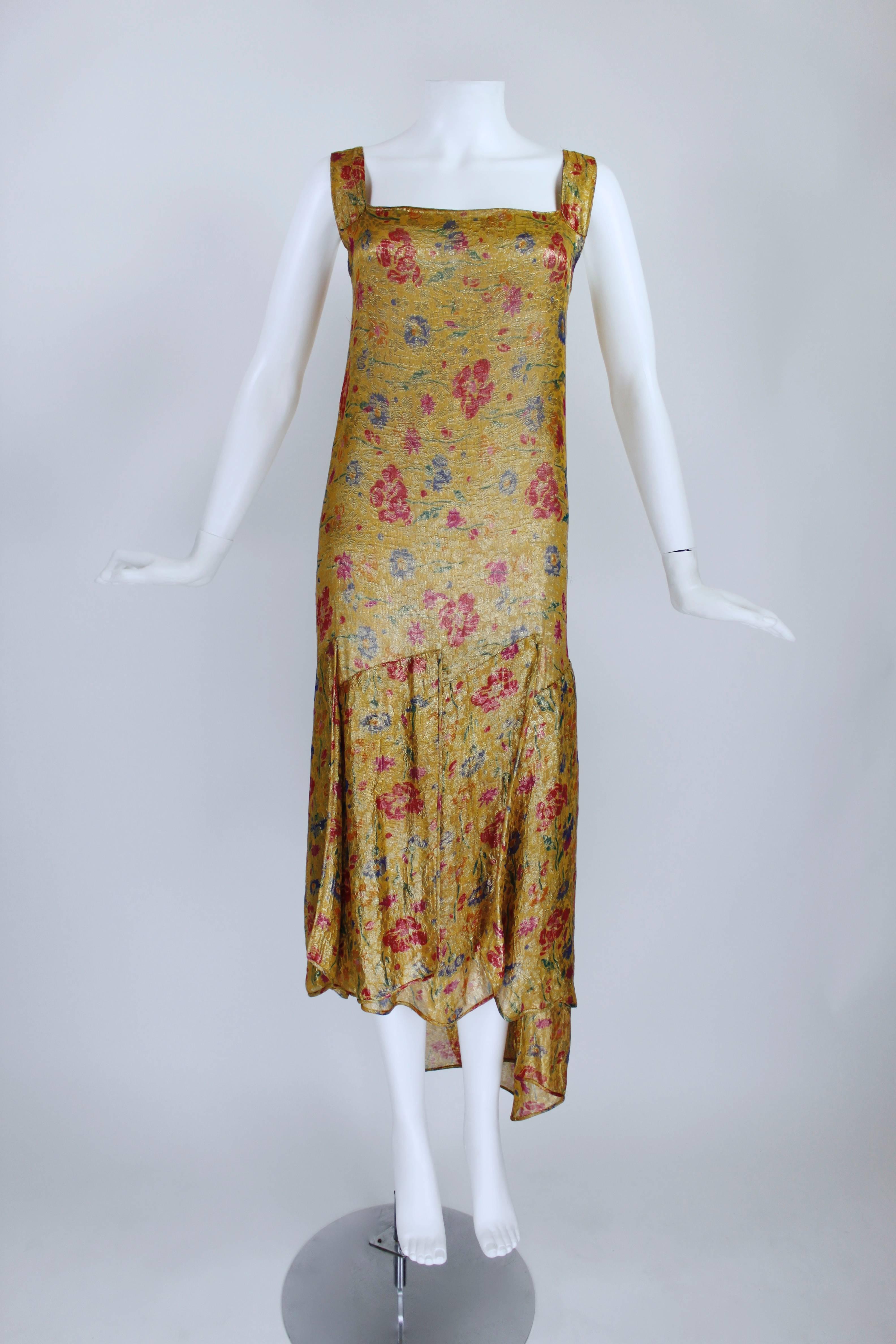 A gorgeous 1920s French lamé cocktail dress, featuring a romantic, vibrant floral pattern laced with metallic gold thread throughout. The silhouette has an iconic drop-waist hem and tiered, asymmetrical panels hanging down--perfect for