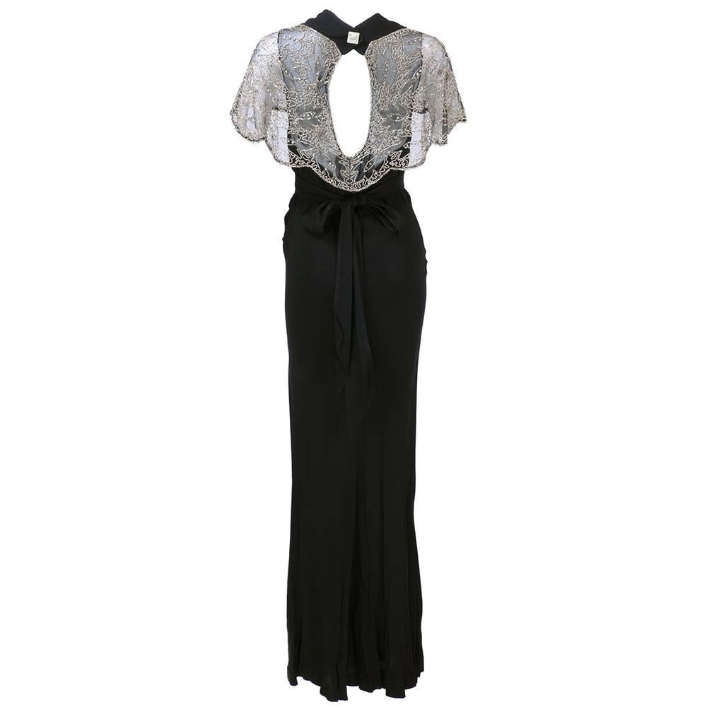 Stunning 1930s gown of bias cut black crepe with attached sheer black net embellished with beads and rhinestones in abstract pattern. Peekaboo back with rhinestone studded deco style button. Snaps at side with attached sash to tie in rear with