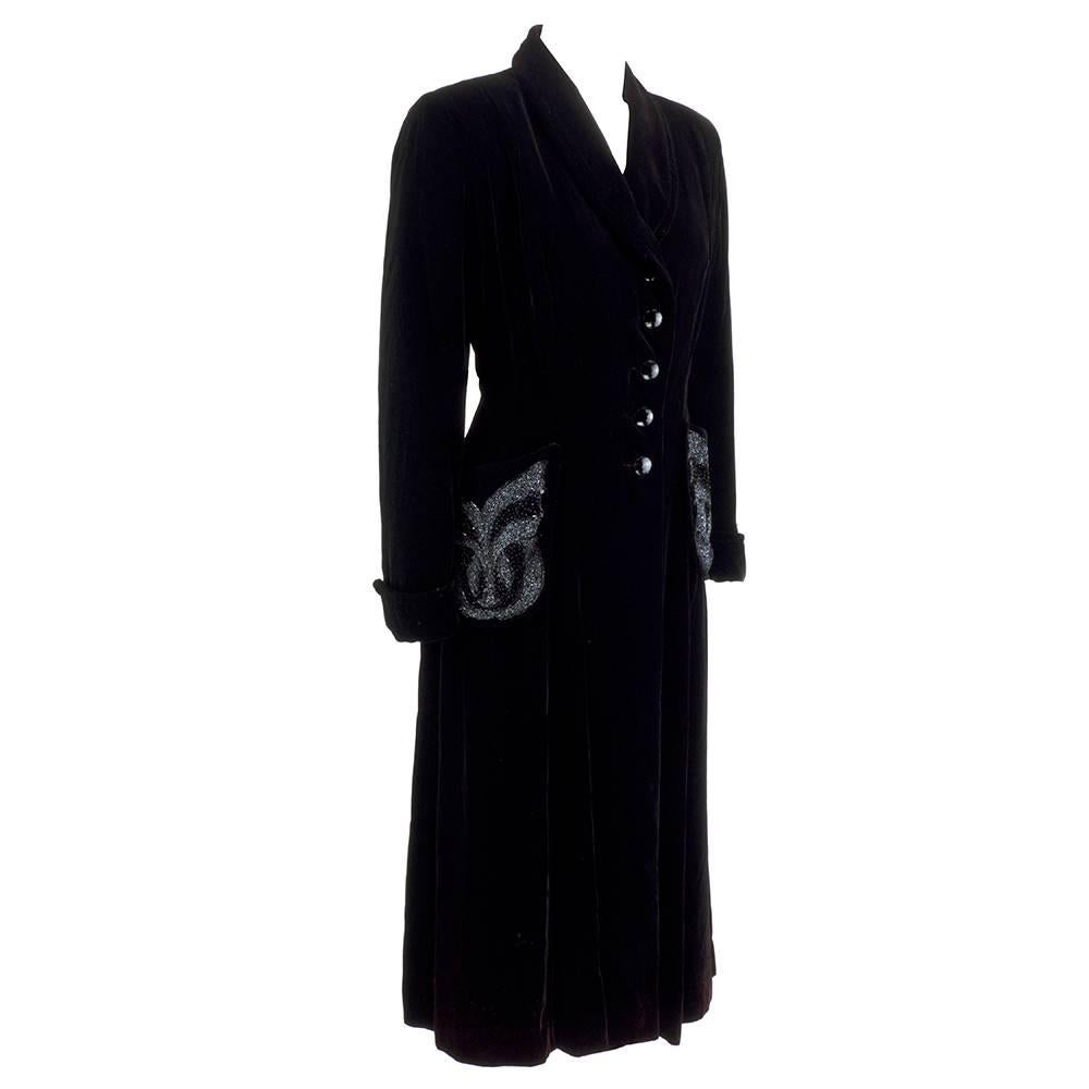 Elegant evening coat circa 1950s. Luscious black velvet in fit and flare style with shawl collar. Fully lined in black satin. Delicate black beading on patch pockets with faceted black buttons down front with cuffed sleeves.  Casts a purple