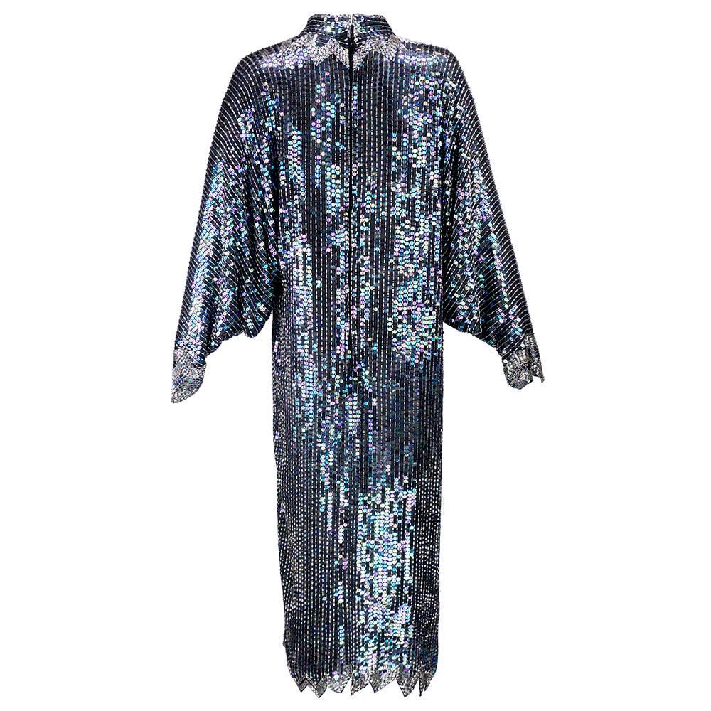 Classic sequined cocktail/evening dress from Halston. Alternating columns of black iridescent sequins and bugle beads cover the dolman-sleeved cocktail dress. Finished with an asymmetric hem bordered in contrasting silver beads.  Backed by silk