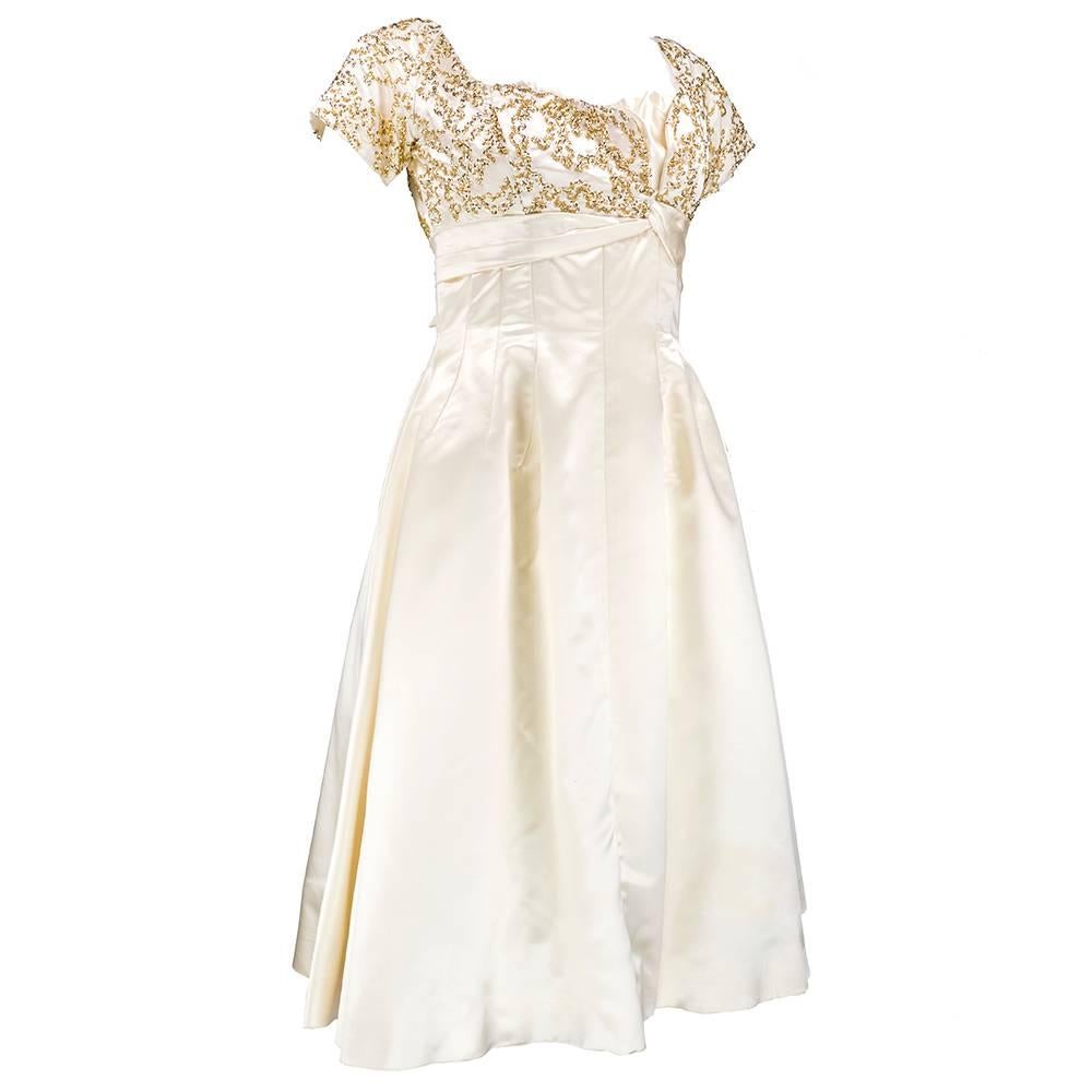 Pretty enough for a princess, Ceil Chapman cocktail dress circa 1950s in a classic fit and flare style. Ivory colored satin with bodice covered in gold tone sequins. Fully boned bodice and built in petticoat. Zips up back with attached bow at rear.