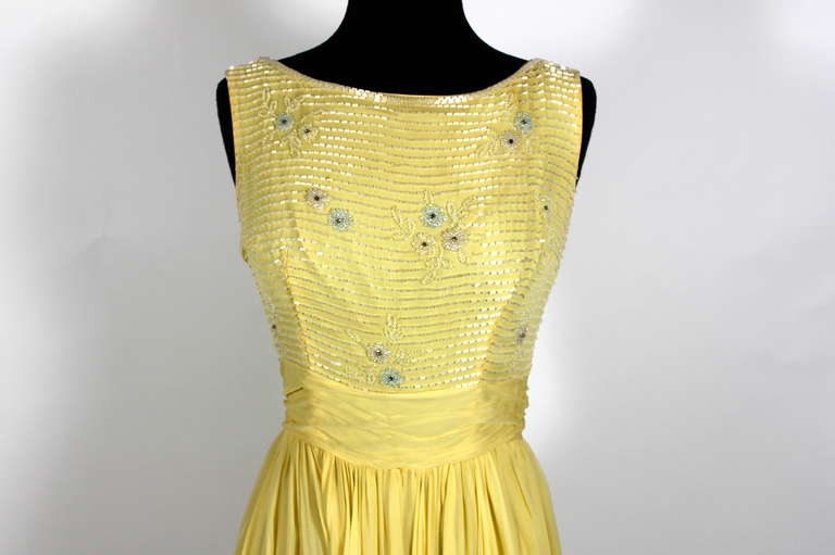 An absolutely lovely layered chiffon party dress from Ceil Chapman. Done in flowing buttercream yellow silk chiffon and embellished with floral motif beading, this is the perfect summer cocktail dress. The tucked chiffon belt attaches in back across
