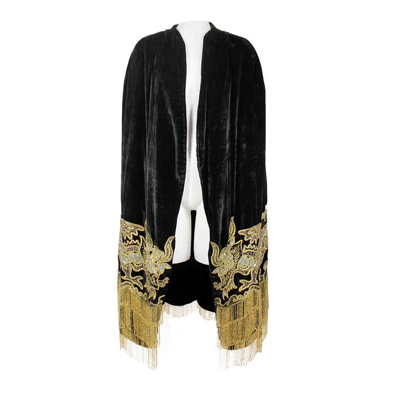 A stunning velvet cape from the 1920s. Done in an ultra luxe black velvet and embellished with gold bullion embroidery, iridescent sequins, and rhinestones forming a dragon motif throughout. The scallop-hemmed cape is trimmed with three tiers of
