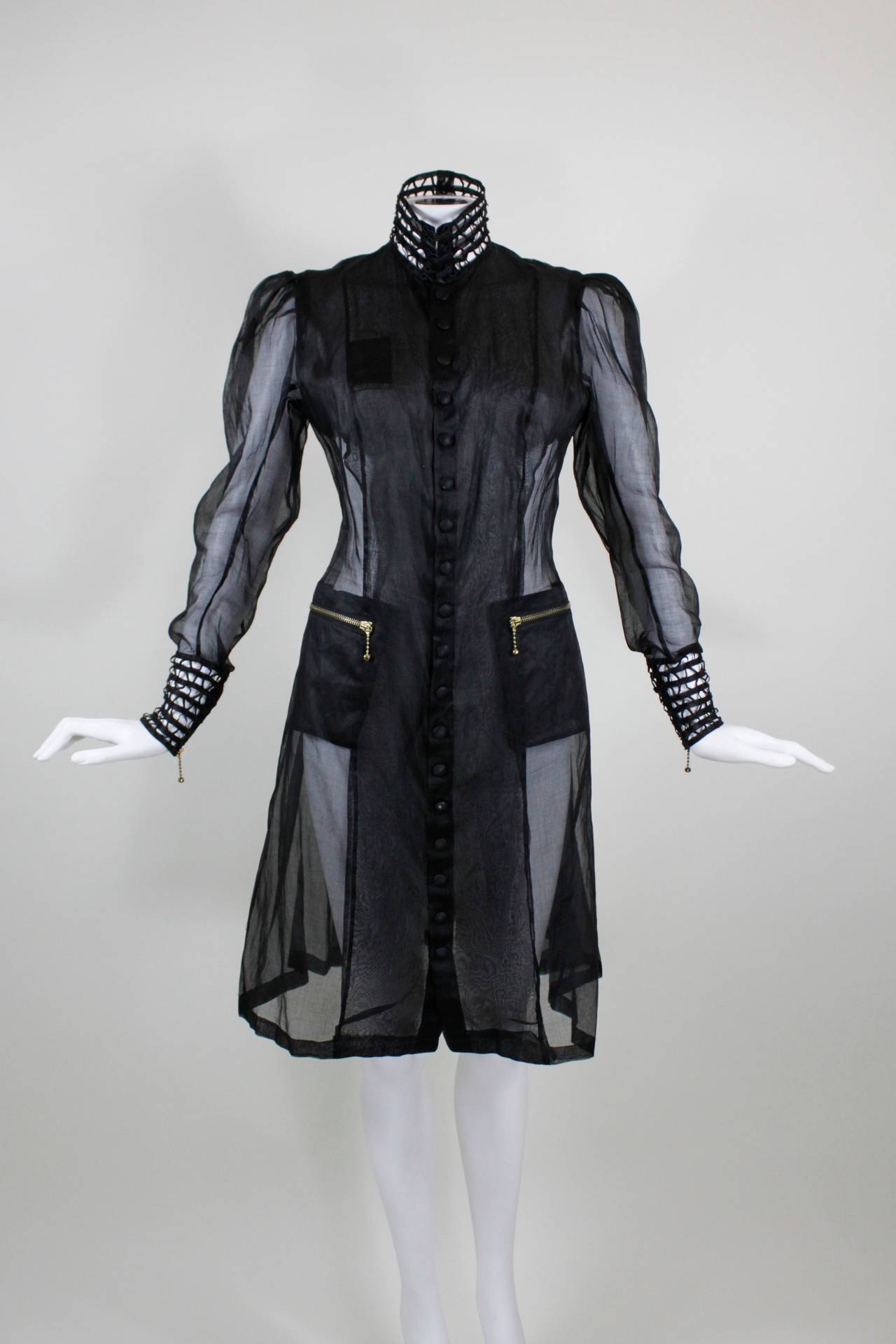 A coat from Jean Paul Gaultier, done in sheer black organza and finished with braided cage cuffs and a high-neck collar. The collar fastens with three hook-and-highs, and the sleeves can be zipped open. The jacket features a snap-front closure, and