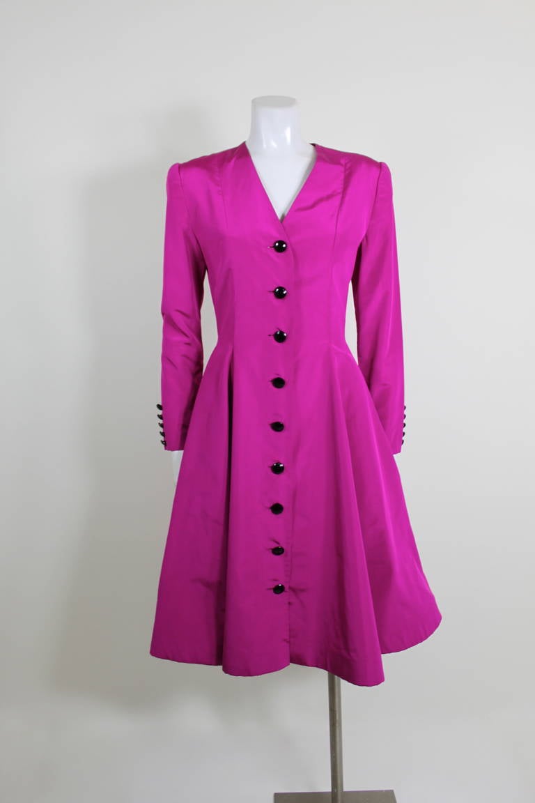 A luxurious, chic, and ultra flattering coat dress from Oscar de La Renta. Done in a beautiful vibrant pink taffeta, the dress features beveled black buttons down the front and on the sleeves. A full, structured skirt accents the nipped waist.