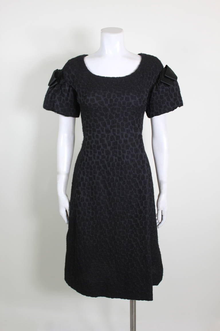 An elegant 1950s cocktail dress from Christian Dior New York. Done in a geometric textured black wool, the dress features a wrap illusion skirt, structured sleeves, and bow accents. 
