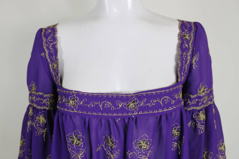 A flowing chiffon peasant dress with a nod to romantic Renaissance silhouettes. Done in vibrant royal purple chiffon and embellished with gold metallic floral embroidery throughout. The dress features a ruffled hem, beautiful lace trim, and buttons