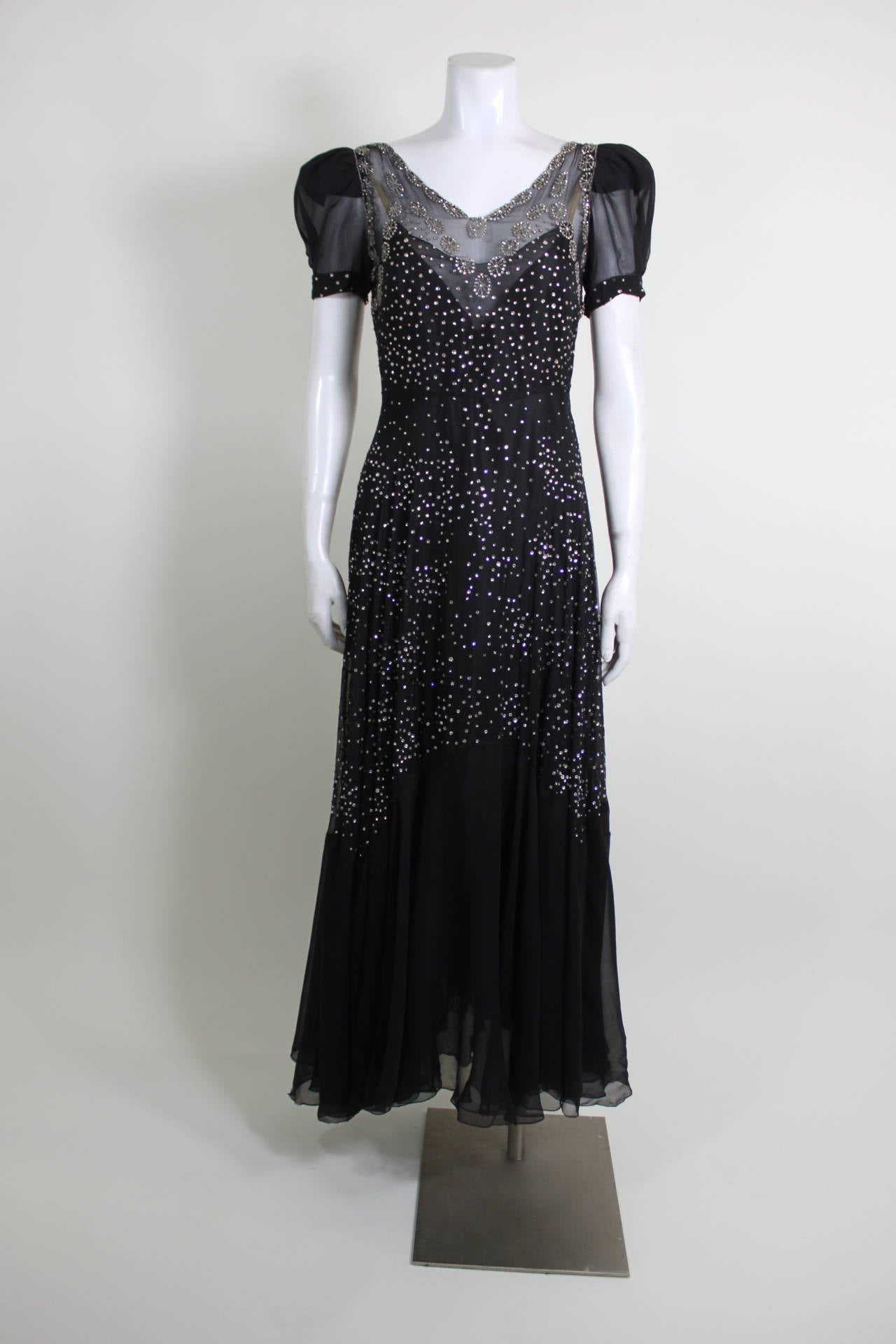 An insanely gorgeous, sparkling bias cut gown from the 1930s. Done in a lightweight black chiffon, the gown is covered in prong-set rhinestones in starburst patterns. Princess sleeves are given a modern silhouette with a chic structured cut. The