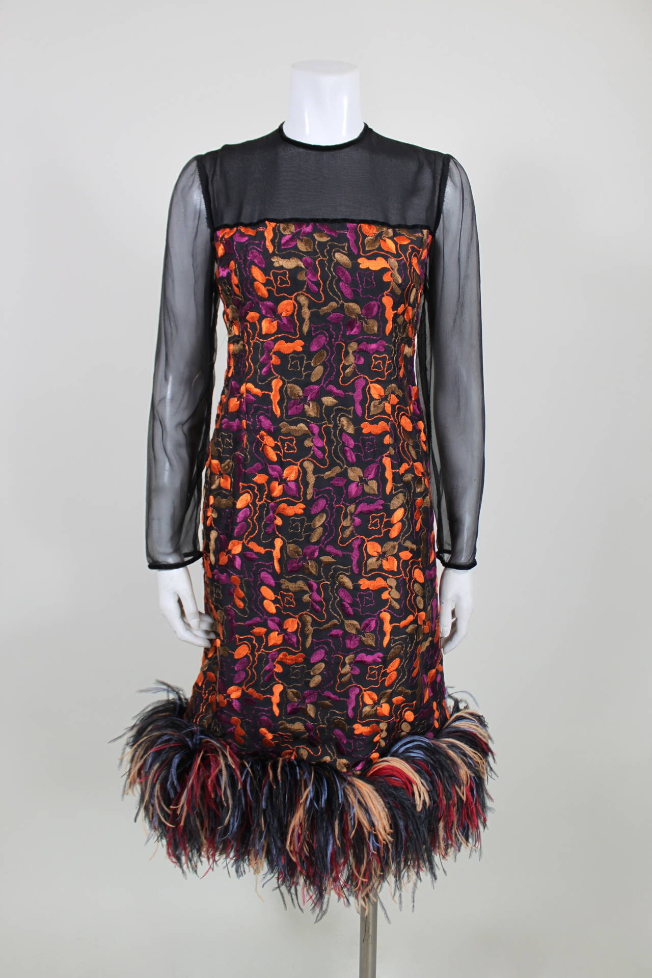 A vibrant, beautifully embroidered cocktail dress featuring a sheer bodice and sleeves. The hem of the dress is trimmed in full, colorful ostrich feathers. Zips and buttons in the back. Fully lined. 
