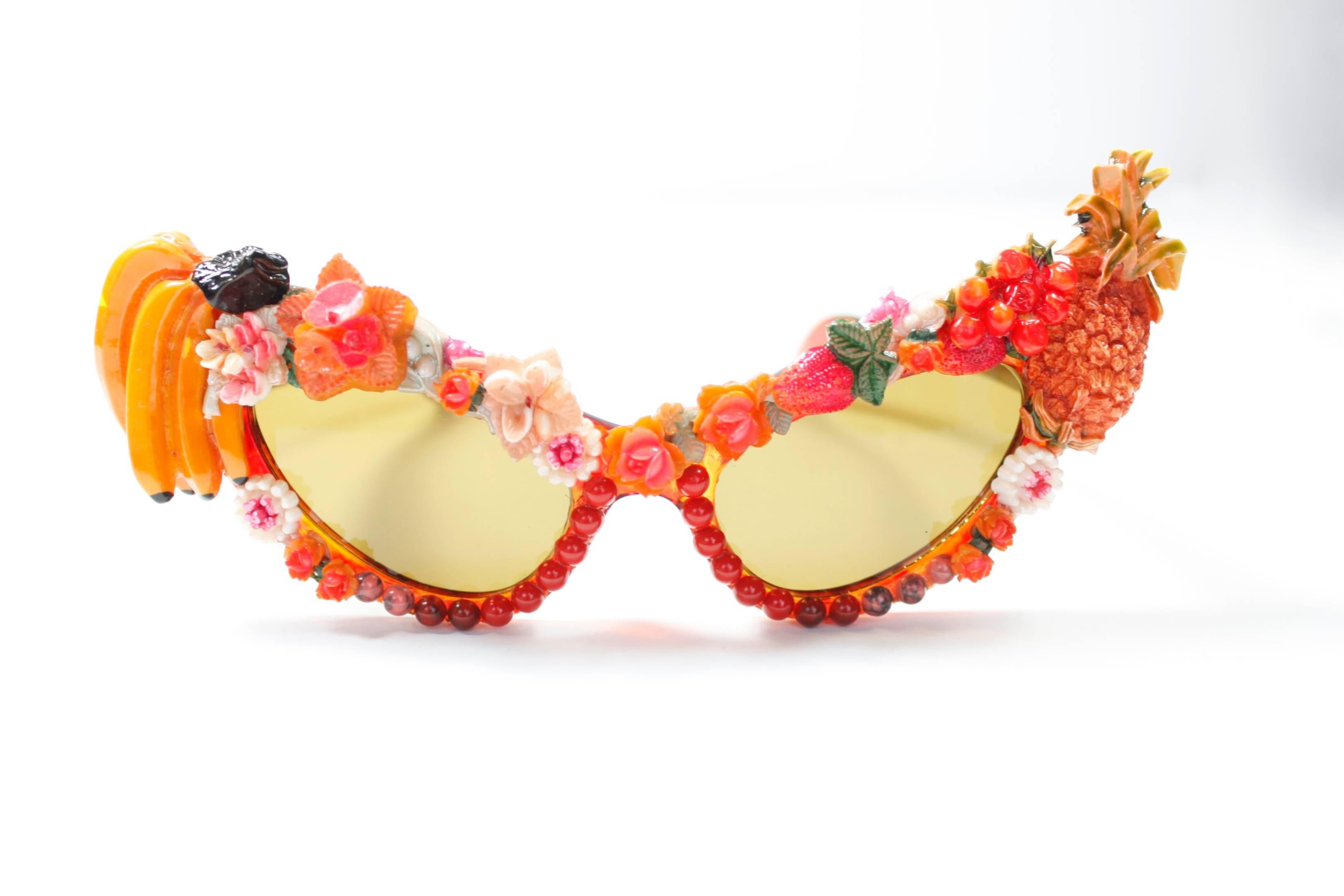 A knock-out pair of statement sunglasses from Mercura Fine Art, featuring an exaggerated cat-eye shape embellished with dimensional, colorful fruit and flowers. The frames are faux-tortoise, and the lens color is a smokey amber. A bright-lit photo