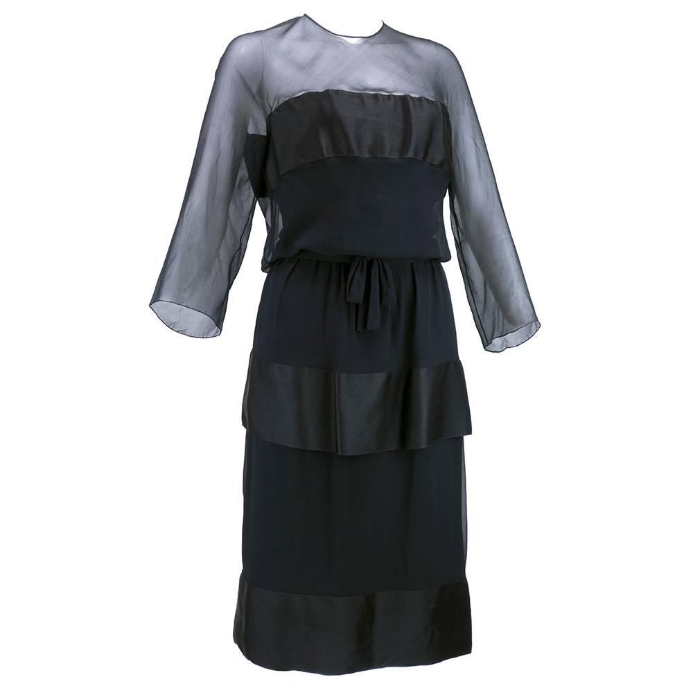 Sheer sophistication. Incredibly chic little black dress by the House of Dior circa late 50s/early 1960s. In chiffon with horizontal bands of satin - blousoned top with quarter sleeves and tiered skirt. Fully lined with boned bodice. Attached belt.