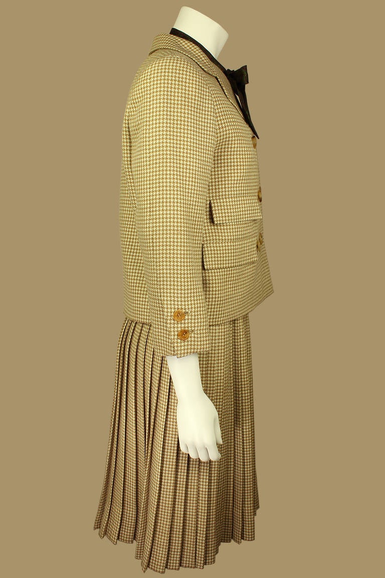 This suit is a classic three-piece Norell with a caramel and creme wool houndtooth three-quarted length sleeve jacket, accordian pleated skirt, and a printed silk blouse. The paisley silk blouse has a bow neck and double-breasted covered buttons.