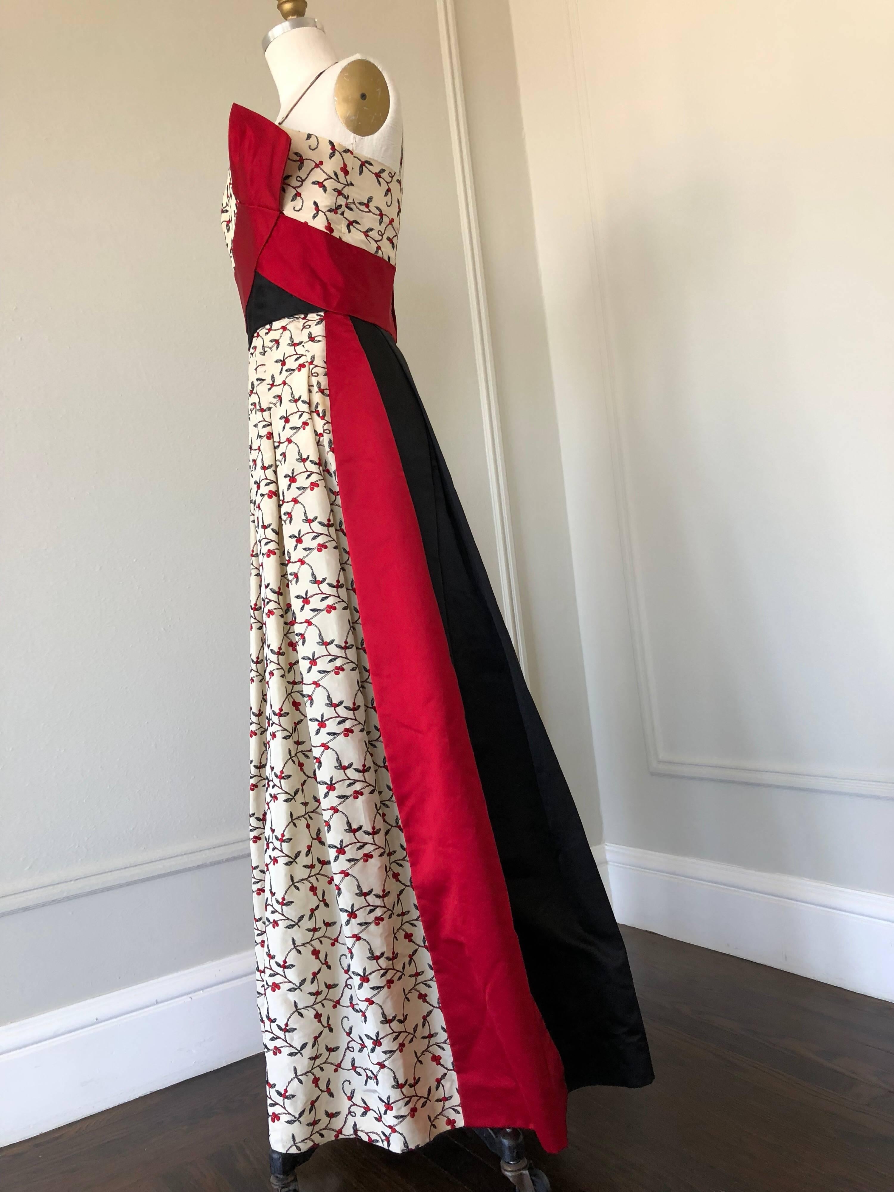 An elegant 1950s evening gown by Oleg Cassini features cream silk embroidered floral motif silk taffeta fabric with a red silk sash at the bodice and black silk satin backside. 
This genius and spontaneous design make this gown modern and stylish in