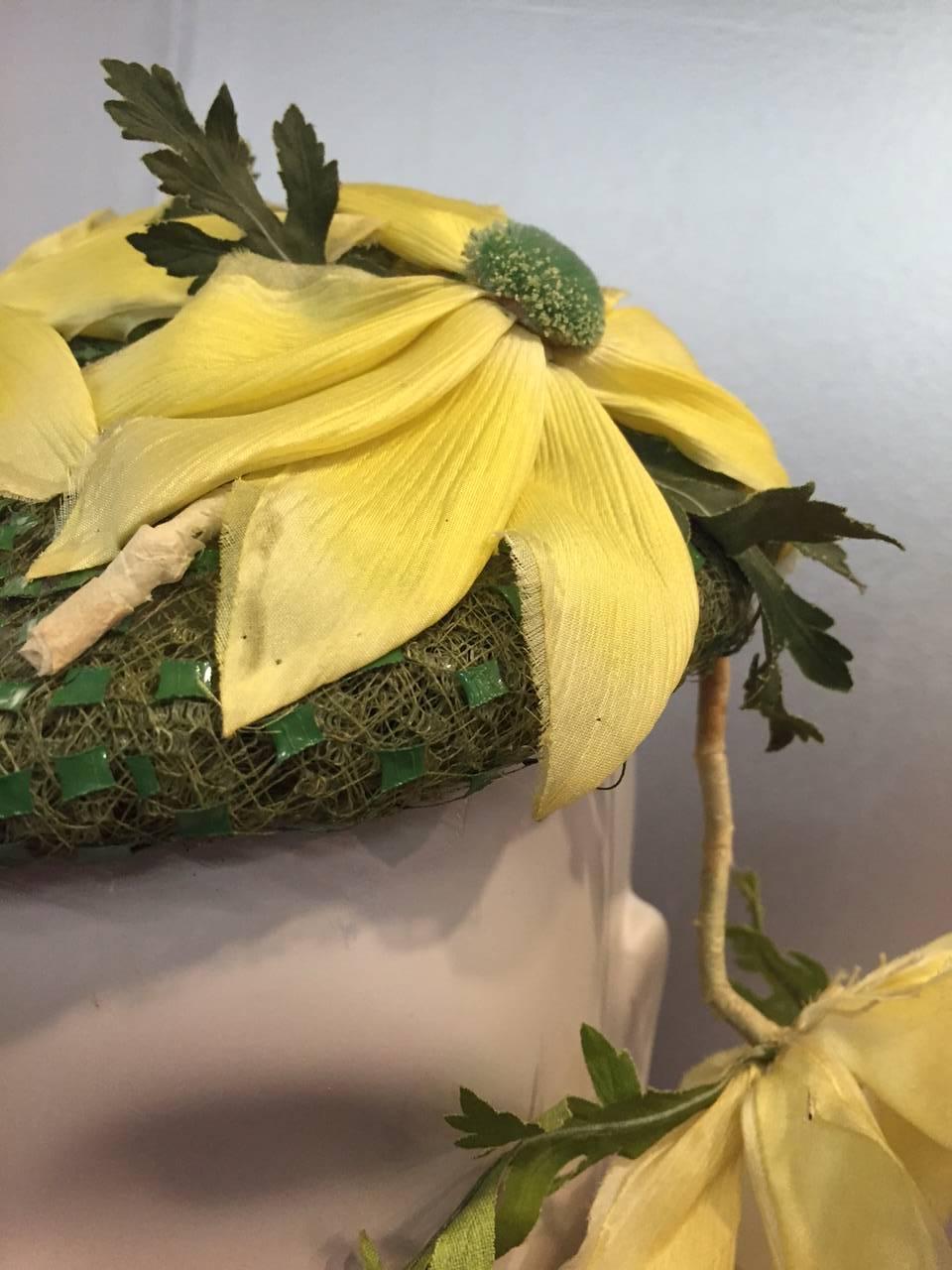 A charming 1950s Saks Fifth Avenue cocktail hat:  green net and veiling topped with beautiful yellow and green silk daisies and one single flower trailing to the side of the face.  Secure wire clamps at sides for a great fit. 