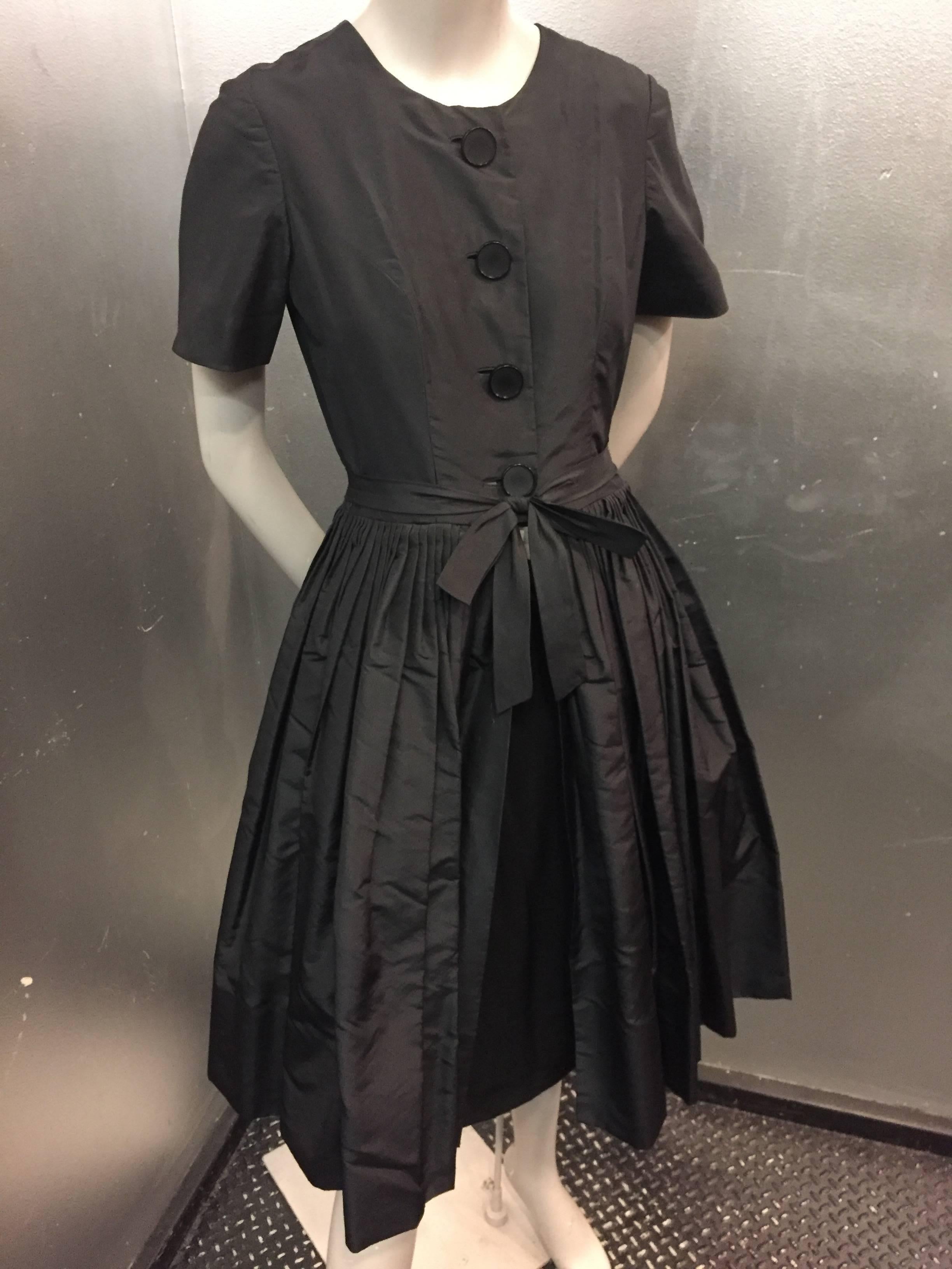 1950s Gustave Tassell black silk taffeta cap-sleeved cocktail dress: Full pleated skirt is open from waist at front to an exposed silk crepe sheath underneath. sheath is lined in silk with side zipper.  Dress buttons at front with a bow at waist. 