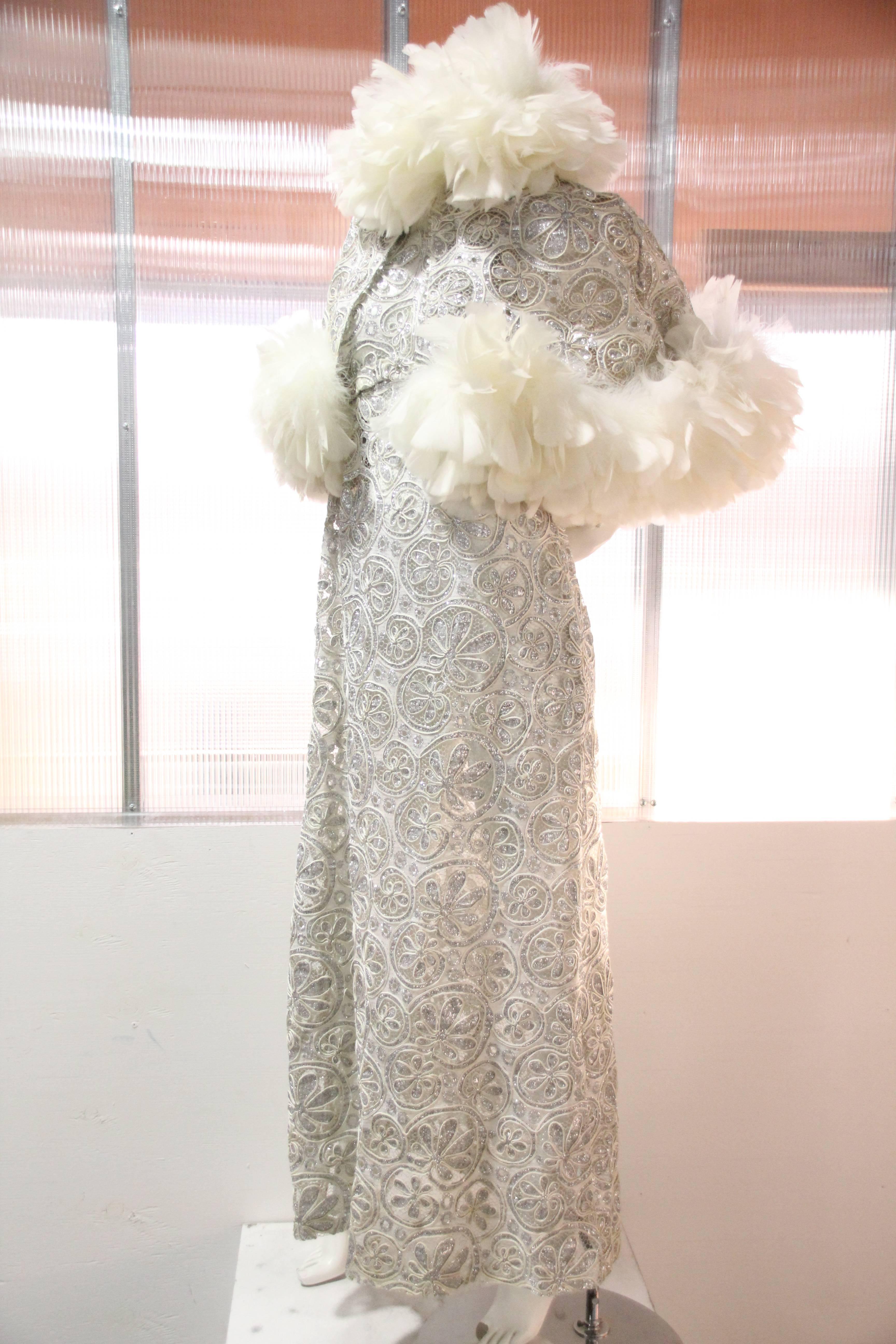 1960s Halter gown of a weighty couched and embroidered silver lame lace: Halter neckline. Collar is trimmed with feather boa and low-cut back is embellished with silver cut-out and eyelet punched leather. Dress is unlined. Caplet us trimmed in white