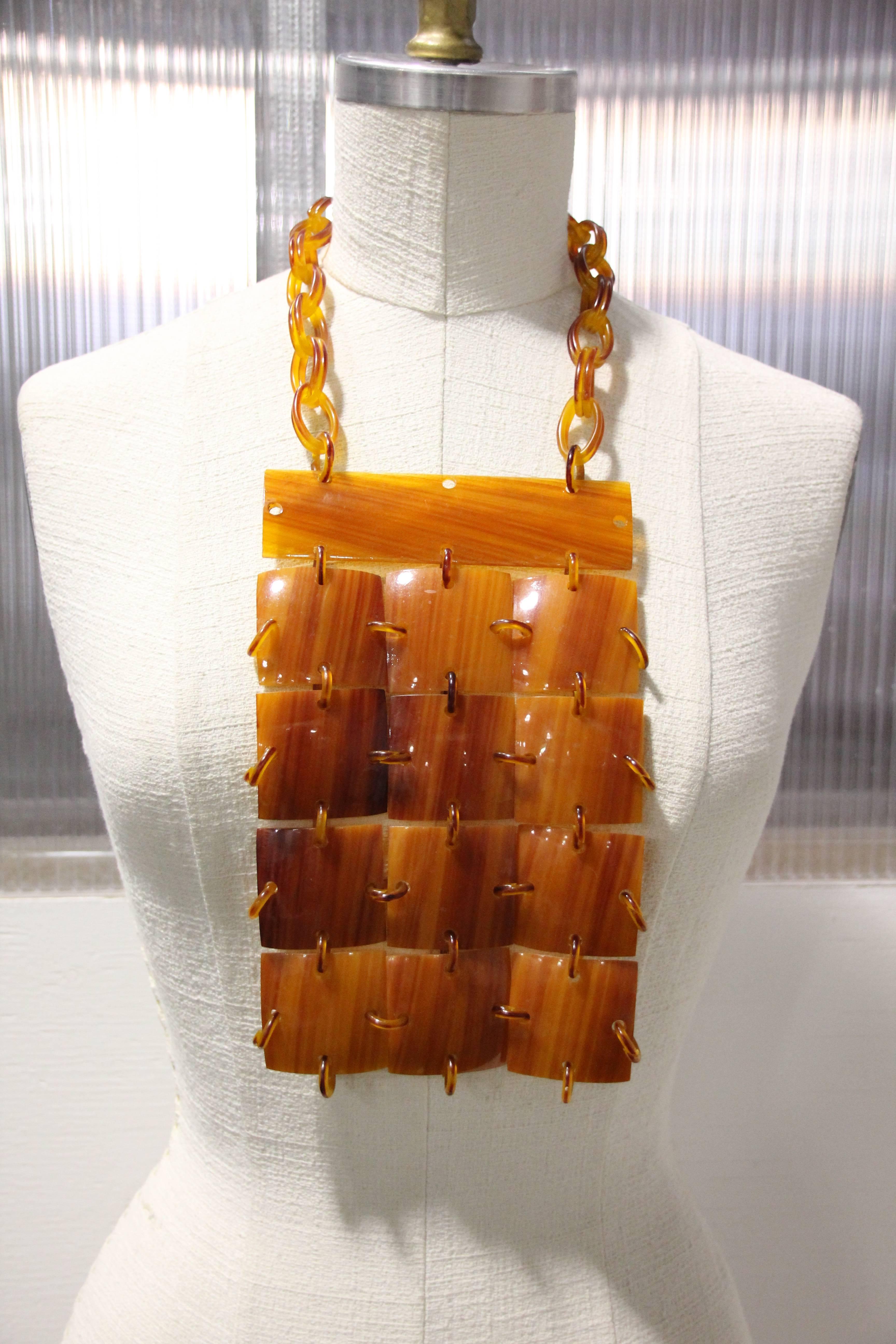 Ce collier plastron en bakélite couleur carmel des années 1960 est composé de carreaux de forme carrée qui sont reliés par des anneaux de sautage du même matériau ainsi que par la chaîne en bakélite autour du cou. 
Une grande barre plate en forme de